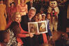 President Clinton and First Lady Hillary Rodham Clinton sit and hold up the book to show the book's illustrations to children sitting around them. People in period costumes can be seen standing in the back ground. a person dressed as a snow man can also be seen in the back ground.