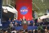 Johnson Space Center President Clinton speaking at Johnson Space Center. Astronauts from NASA mission STS-95 stand behind the President as he delivers his remarks. A red flag with the NASA emblem and two space shuttles are also in the background