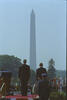 President Clinton and President Václav Havel of the Czech Republic stand on the dais on the South Lawn of the White House looking toward the Washington Monument. Trees can also be see in the background. A man in a uniform is below the dias, also looking toward the Washington Monument (an obelisk)