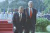 President Clinton and President Václav Havel of the Czech Republic walk together in the South Lawn of the White House. Havel looks down and Clinton looks up.  A red carpet, american armed forces troops, flags, green trees, and greenery can be seen in the background.