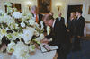 President Václav Havel signs a book with white pages. A pen box sits on the open guest book. The book sits on a round table with a white table cloth. A large flower arrangement of white flowers sits on the table. President Clinton stands looking down at President Havel as he signs the guest book. First Lady Hillary Rodham Clinton and President Havel's wife Dagmar Havlova can be seen standing in the room. Two unidentified men stand in the background.