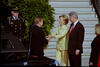 First Lady Hillary Rodham Clinton stands greeting President Václav Havel. First Lady Hillary Rodham Clinton extends both hands towards Havel. She wears a yellow pantsuit. President Clinton stands looking on. President Havel stands beside a black car. A uinformed soldire stands in the background. A green bush and stairs are also in the background. An unknow woman stands lookiing toward the the Clintons and Havel.