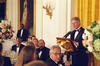 President Clinton stands in a tuxedo behind a gilded eagle lectern. Guests to a state dinner sit around a table, looking up at President Clinton. The President Stands in fron of a gold colored curtain.