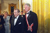 President Clinton wears a red and white sash over black tuxedo suit and white shirt.  President Clinton stands, posing for a photograph with President Václav Havel. First Lady Hillary Rodham Clinton stands, clapping and smilling behind the two presidents.