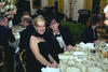 Director of the Office of Scheduling, Stephanie Streett poses for a photograph with musician, Ric Ocasek, at the White House State Dinner. Both sit at a round table. Gold rimmed White House Plates and table ware sit on the table. Ocasek wears a black tux with a white shirt. Street wears a black dress and black scarf. 