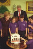 President Clinton and First Lady Hillary Rodham Clinton stand behind children as one child sits lighting a menorah. The children wear purple polo shirts. 