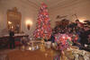 First Lady Hillary Rodham Clinton stands in a room with a Christmas tree made of imitation fruit. The tree and other decorations in the room are pink, violet, and green. A  group of press point cameras and microphones toward the First Lady.