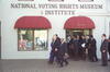 President Clinton, Correta Scott King, and Rep. John Lewis walk in front of the National Voting Rights Museum and Institute. A group of people stand behind Clinton, King, and Lewis. National Voting Rights building is white with red awnings over the window and door.