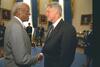 President Clinton stands shanking ands and looking directly into the eyes of John Hope Franklin. Franking wears glasses. Both men wear suits. 4 other people can be seen standing in the background of the photograph.