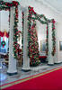 White columns covered in lighted garland with red bows. A lit Christmas tree can also be seen in the photograph.