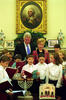 President Clinton and First Lady Hillary Rodham Clinton stand behind a group of Children. The children look at books and sing. A lit menorah sits on a table in front of the children
