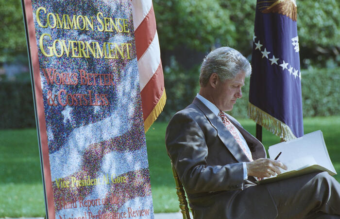 President Bill Clinton seated outdoors, writing in a binder. Clinton sits in profile facing right, wearing a gray suit and a patterned red tie. He is focused on a large open binder resting on his lap, holding a pen in his right hand. To his left, a large colorful vertical banner features a star-spangled design and reads "COMMON SENSE GOVERNMENT" in gold lettering, with the subtitle "Works Better & Costs Less.
