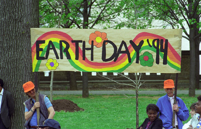 Two Men in orange hats hold an "Earth Day '94" hand painted sign. the sign is brightly pained with flowers and a rainbow.