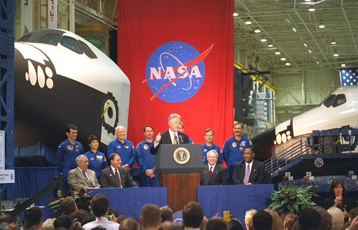 President Clinton speaking at Johnson Space Center. Astronauts from NASA mission STS-95 stand behind the President as he delivers his remarks. A red flag with the NASA emblem and two space shuttles are also in the background