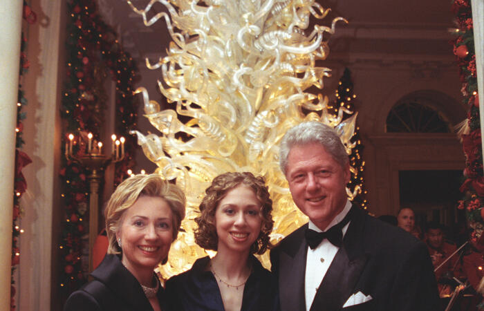President Clinton, Hillary Rodham Clinton, and Chelsea Clinton stand in front of a Dale Chihuly Christmas Tree in the Grand Foyer of the White House. The tree is lit brightly. 