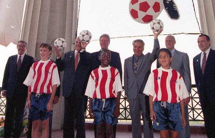 A wide-angle, medium shot features a group of individuals posing together on an indoor balcony against a net backdrop with a soccer ball decoration. Standing in the front row, from left to right, are three children, each wearing a white soccer jersey with vertical red stripes and dark blue shorts. Behind them are several adults in business suits, including Bill Clinton, who is positioned in the center, and others holding soccer balls.