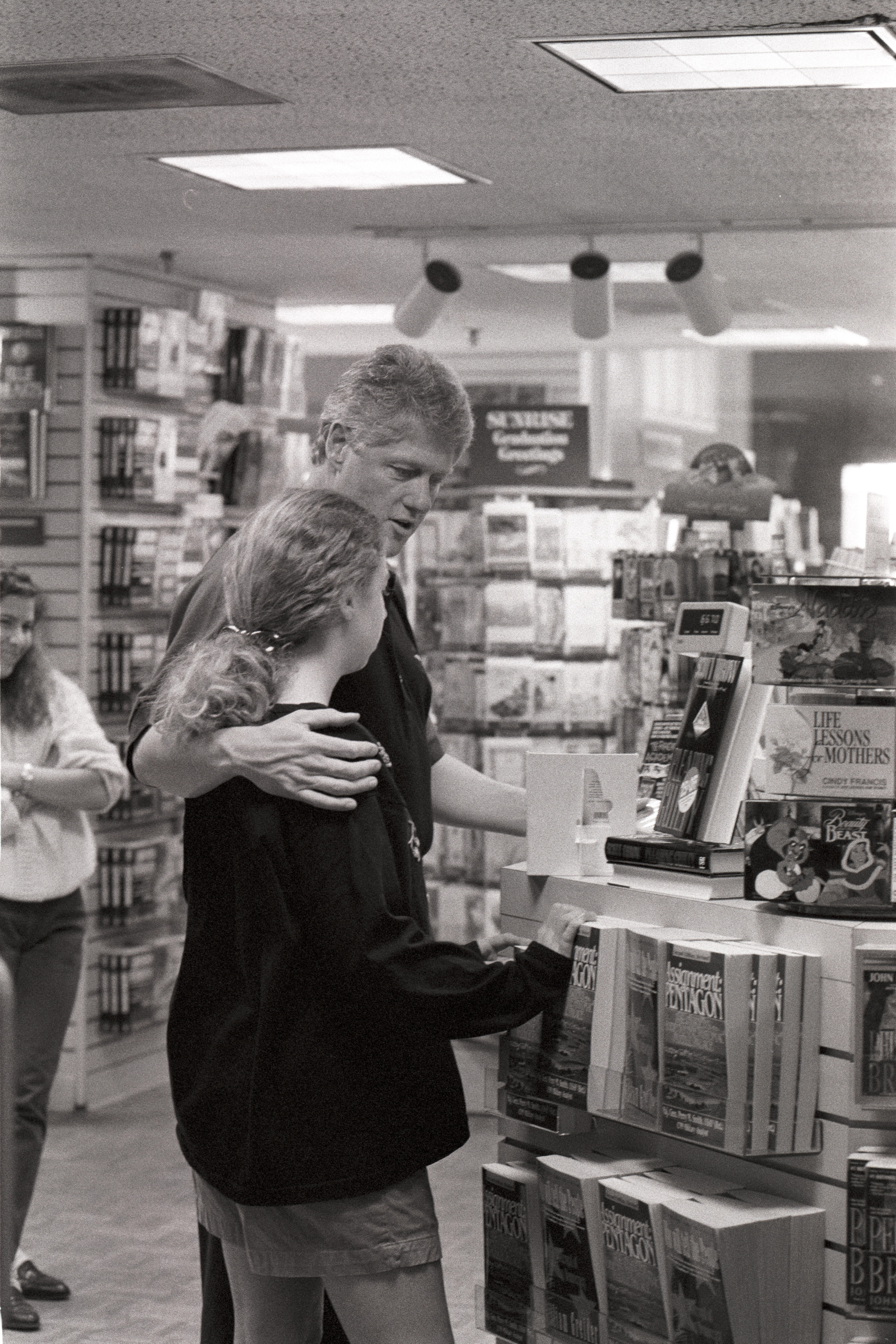 Black and white photograph. President Clinton stands with his arm around his daughter, Chelsea Clinton, while looking at books