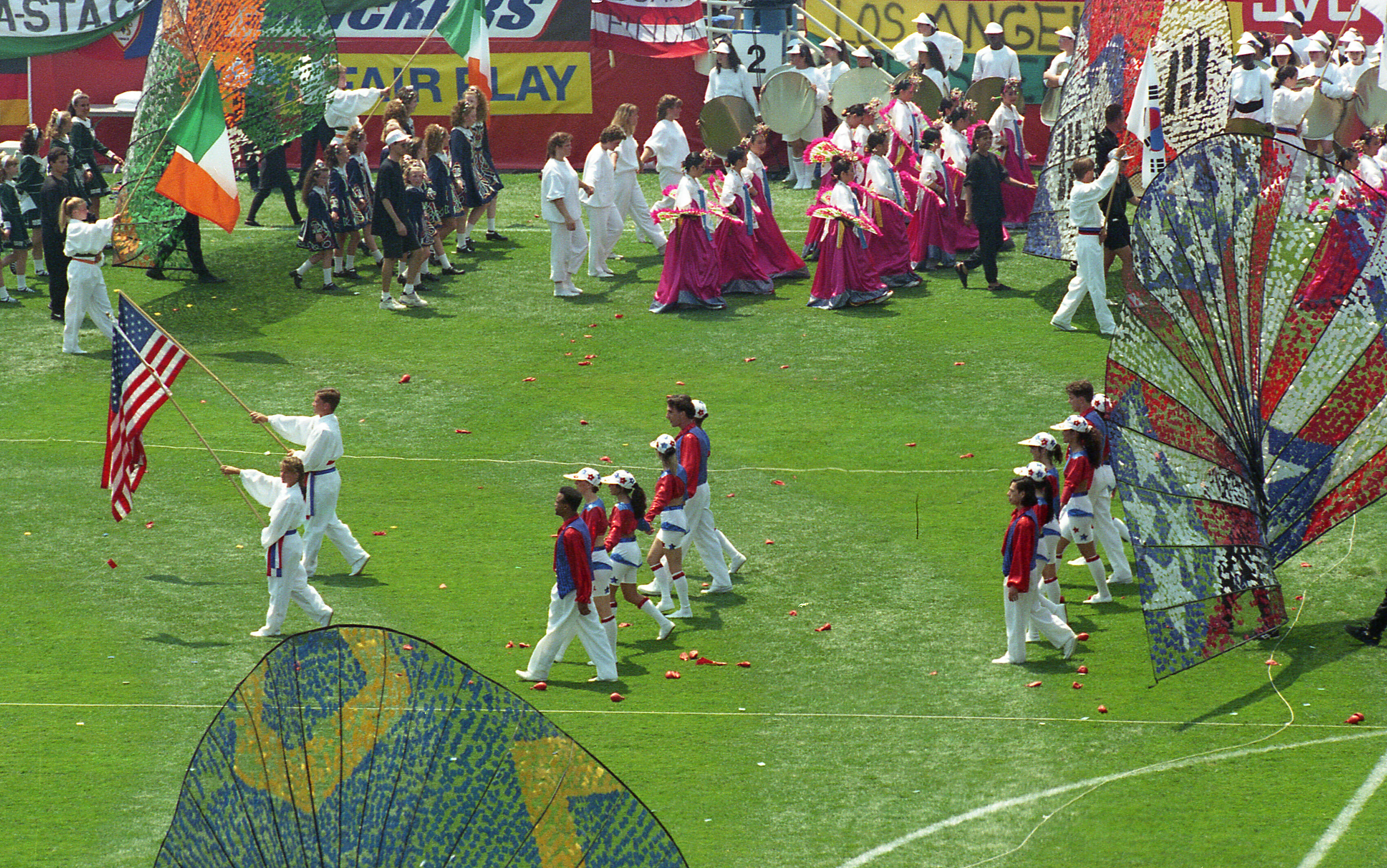 An overhead shot shows the World Cup opening ceremony on a green field. People in colorful costumes, including white, red, and blue outfits, march across the grass. Some carry large flags, like the American and Irish flags, while others hold ornate, fan-like props. In the background, a banner reads "FAIR PLAY" next to more flags and spectators, creating a vibrant, celebratory atmosphere.