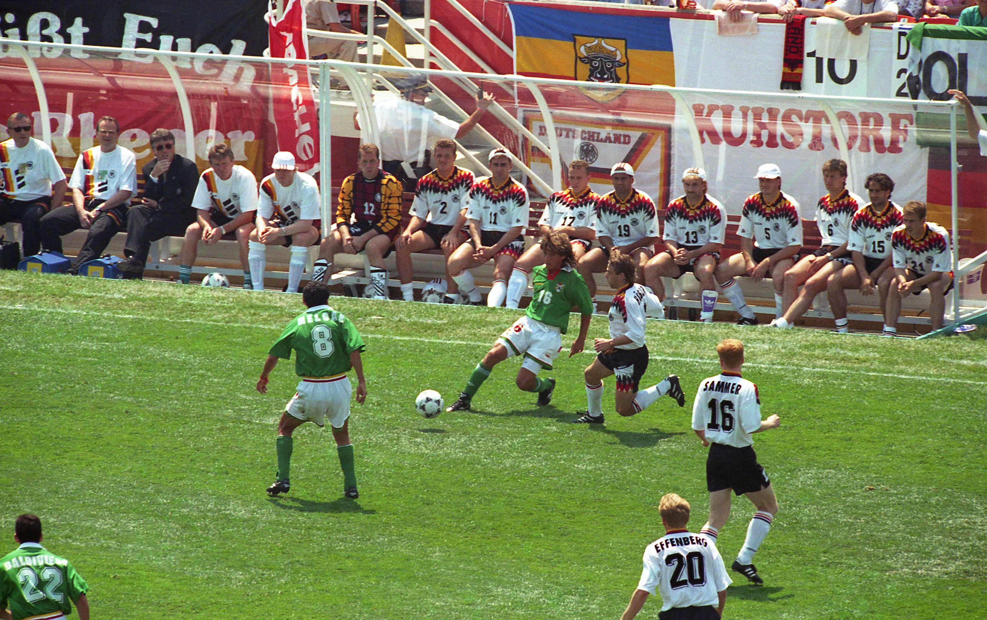 An action shot from a soccer match shows a player in a green jersey (number 16) controlling the ball on a sunny grass field. He is pursued by German players in white jerseys and black shorts, including (number 16) and (number 20). In the background, the German substitute bench sits in a transparent dugout, with various banners and a German flag visible behind them.