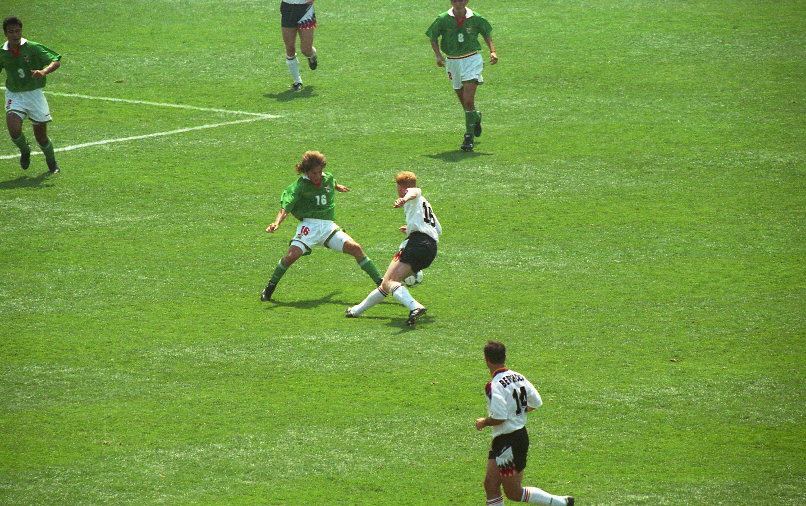 In this action shot from an international soccer match, a German player in a white jersey and black shorts (number 16), battles for the ball on a sunny grass field. He is closely marked by an opposing player in a green jersey, likely from Bolivia, who also wears the number 16. In the foreground, another German player (number 14), runs toward the play, while other players in green jerseys are visible in the background.