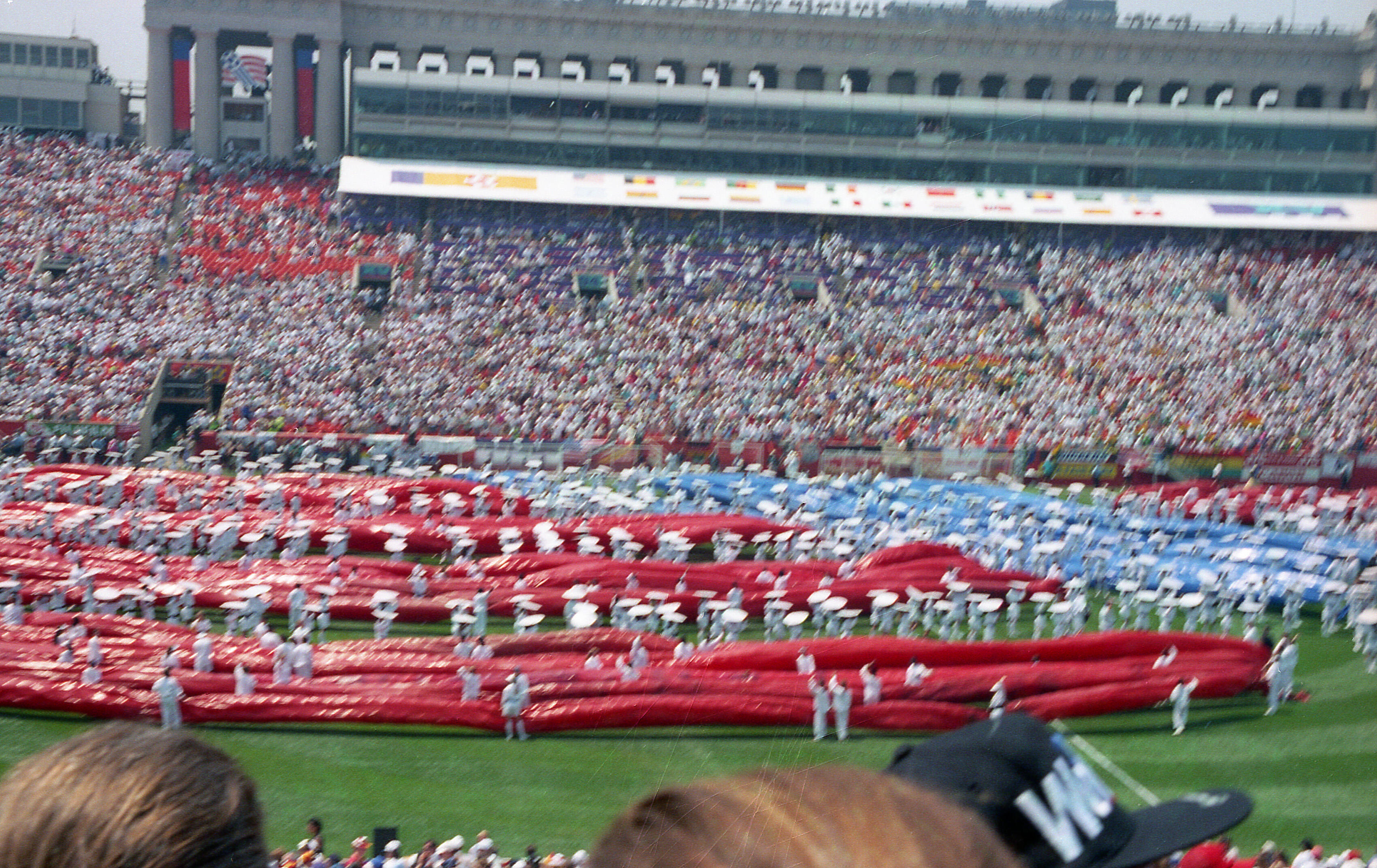 An eye-level shot captures a massive crowd at a stadium, where a sprawling red and blue fabric display covers much of the field. Dozens of people in white uniforms stand amidst the fabric, holding white poles that appear to support the display. In the background, the stadium stands are packed with spectators, and a large multi-story building with flags on top rises behind the seating area. The overall scene is bright.