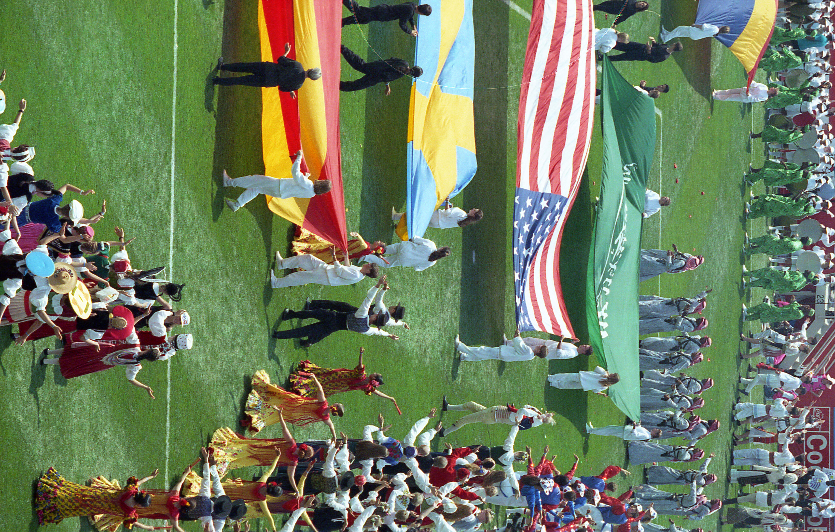 This high-angle photograph captures a segment of the opening ceremony for the 1994 FIFA World Cup at Soldier Field. On the bright green grass, various groups of performers in cultural attire celebrate global diversity. Several groups hold large, horizontal flags aloft, including the flag of the United States and the flag of Saudi Arabia. Other performers are dressed in vibrant, traditional costumes, such as flamenco-style dresses and folk outfits, dancing in choreographed lines. 