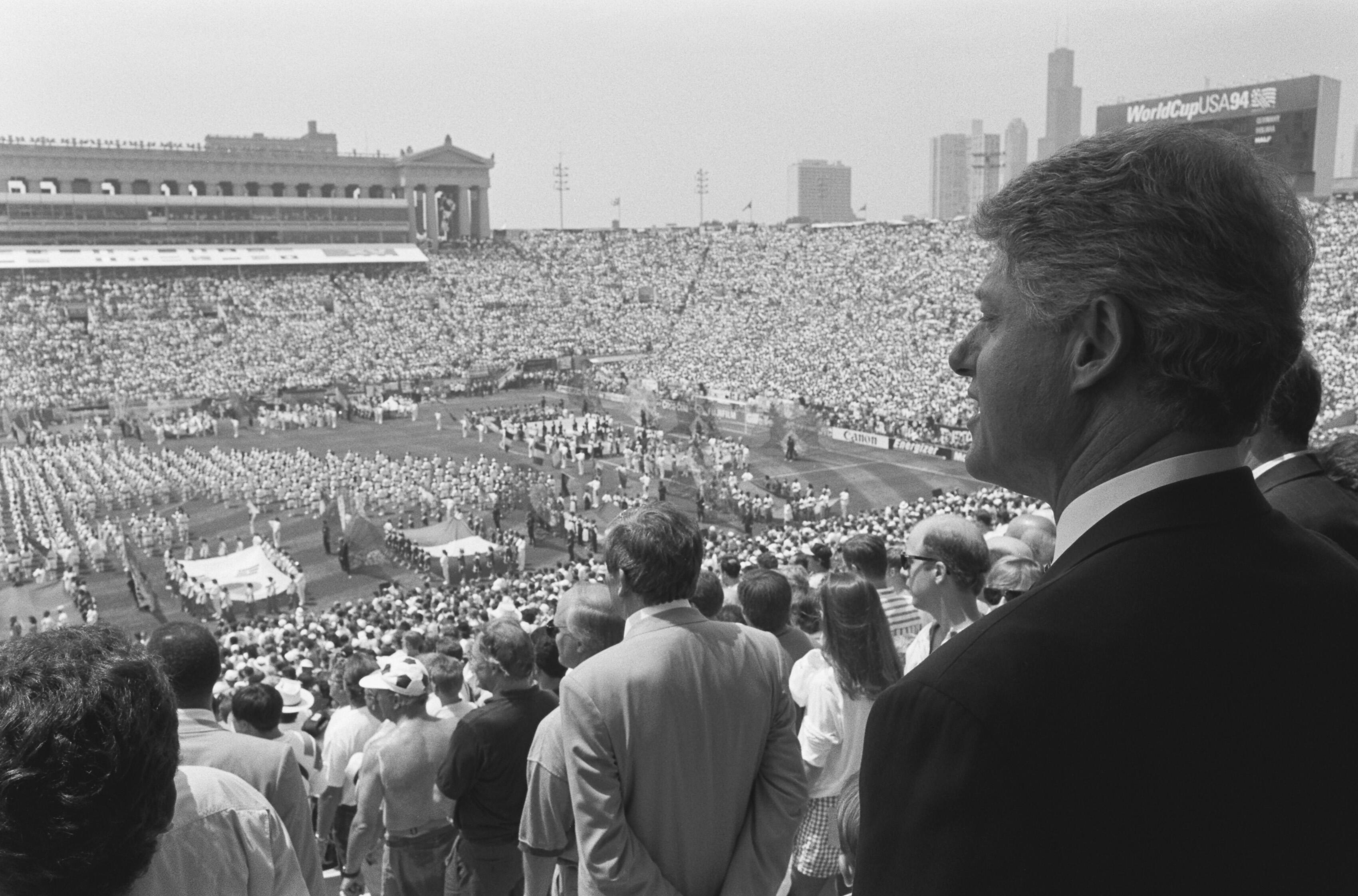 This black-and-white photograph shows President Bill Clinton in profile, overlooking the 1994 FIFA World Cup opening ceremony at Soldier Field. In the foreground, Clinton stands in a dark suit, his gaze directed toward the field where performers are arranged in large formations. The stadium's stands are overflowing with spectators, and in the distance, a large electronic scoreboard displays the text "WorldCupUSA94." The Chicago skyline, including the Willis Tower (then the Sears Tower), is visible.