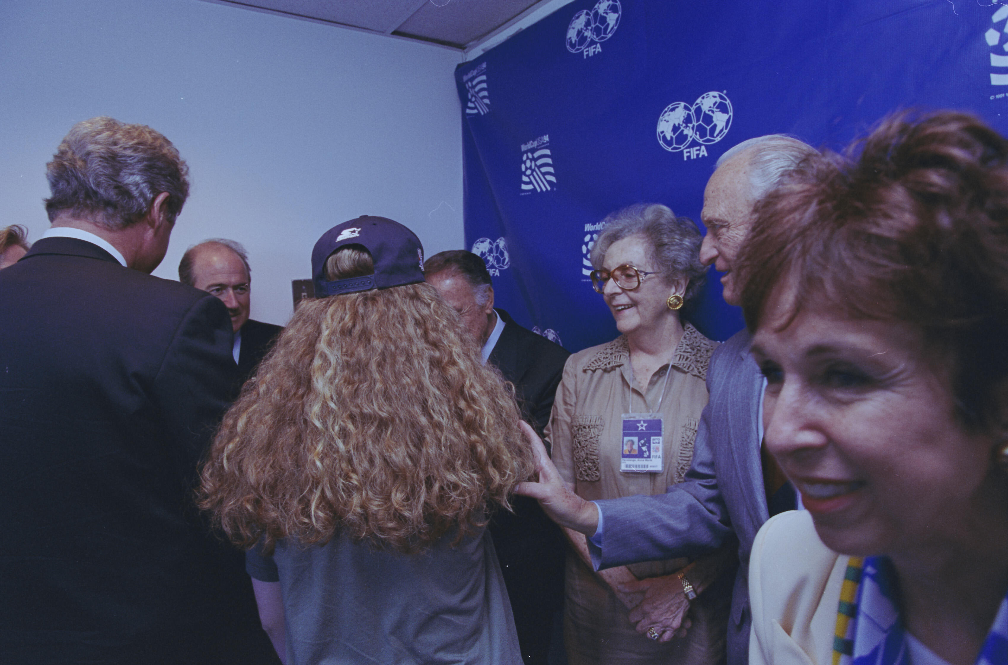 In the center, a person (Chelsea Clinton) with long, curly blonde hair wearing a purple baseball cap stands with their back to the camera. To their right, an older woman with short blonde hair, wearing glasses and a tan blouse with a prominent event credential, smiles toward someone off-camera. Several men in suits are partially visible in the background and foreground, including Bill Clinton, whose back is to the camera on the left. The lighting is slightly dim, typical of an indoor event space.