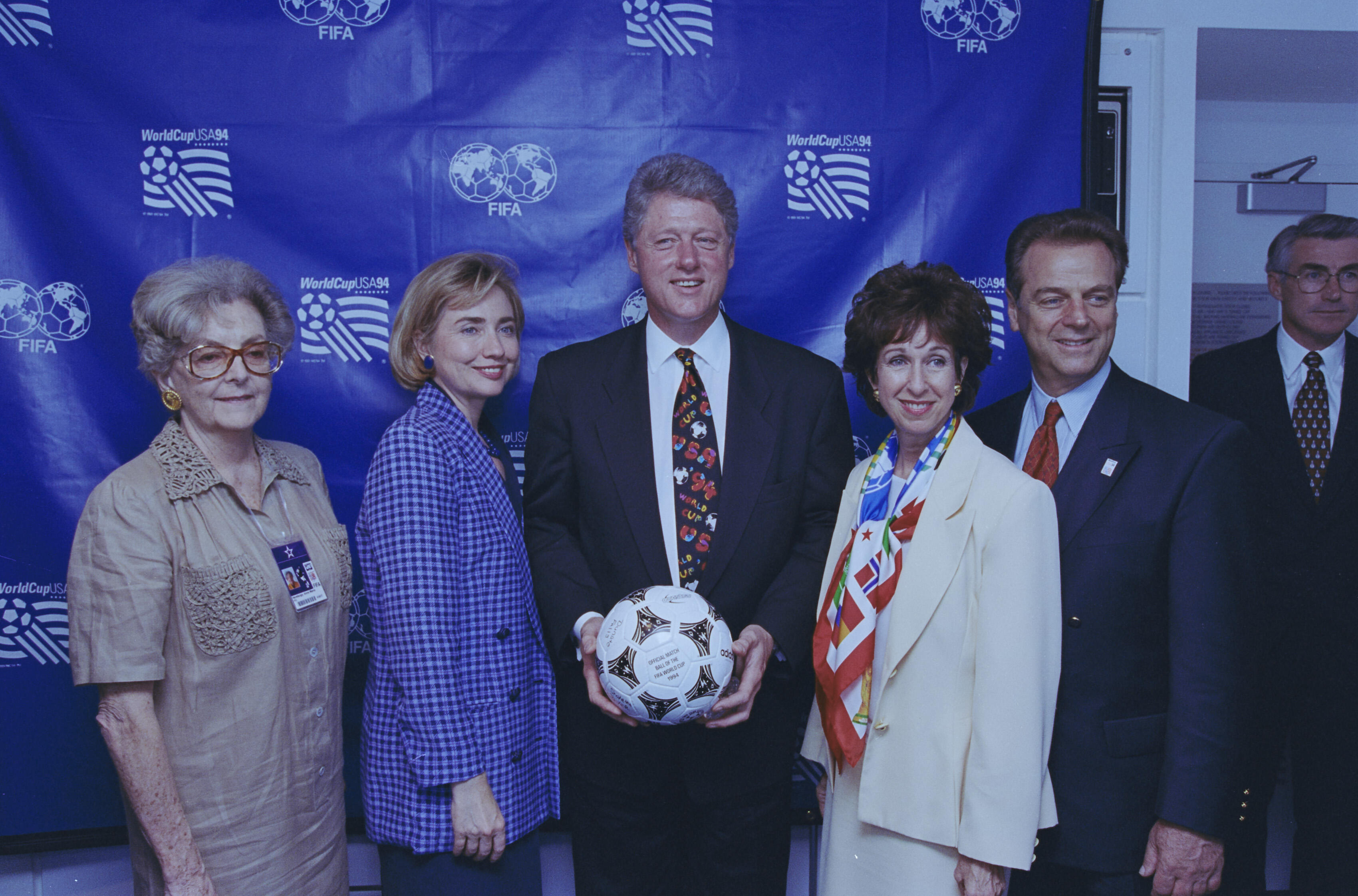 In the center, President Clinton holds the Questra, the official match ball of the tournament. To his left, Hillary Clinton wears a blue checkered blazer, and to his right is Alan Rothenberg. Also present are two women: one on the far left wearing a tan blouse and an event credential, and another in a cream-colored blazer with a colorful scarf standing next to the President. They are all positioned in front of a blue step and repeat backdrop featuring the WorldCupUSA94 and FIFA logos.