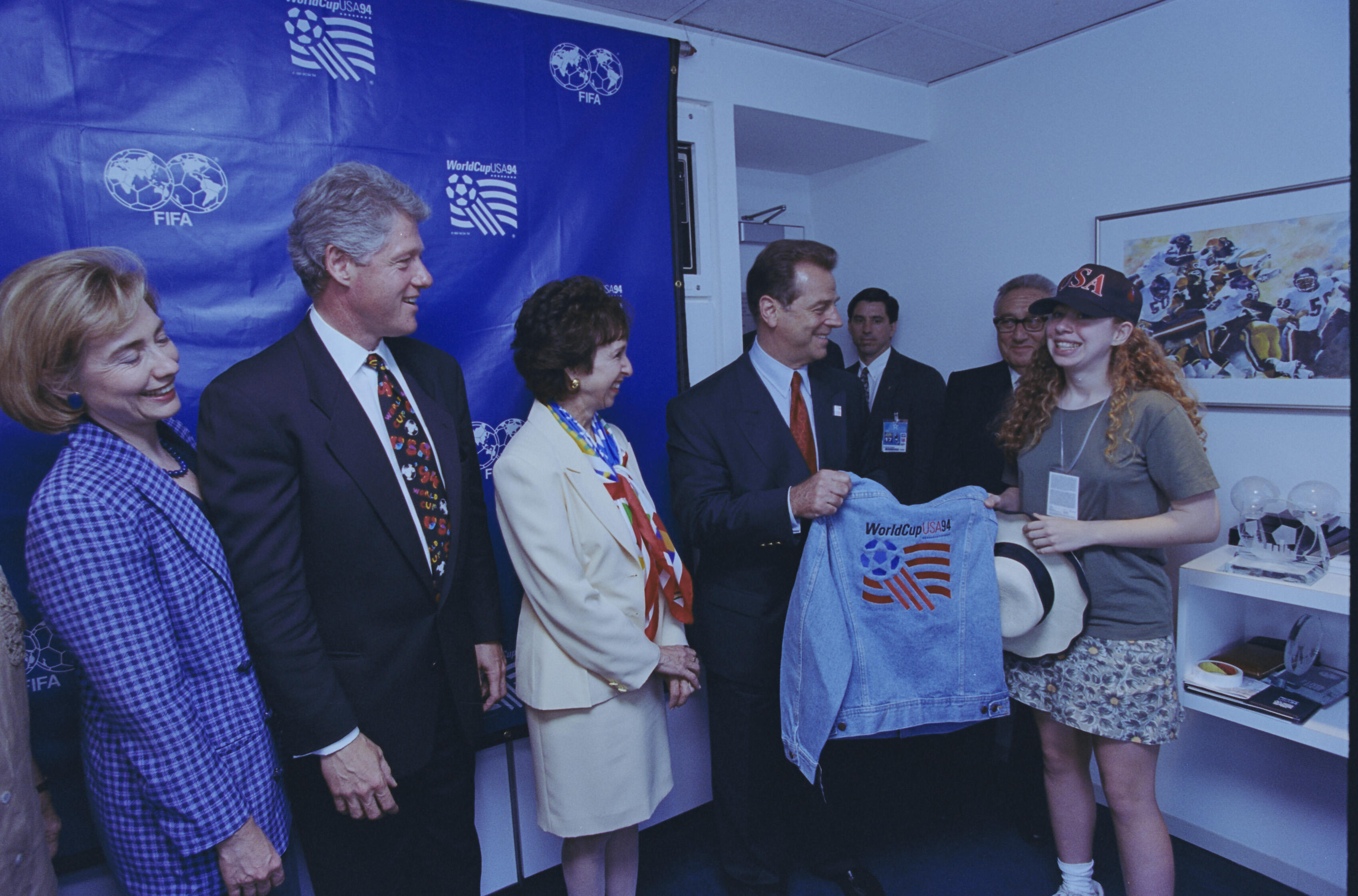 On the left, First Lady Hillary Clinton and President Bill Clinton look on with smiles as Alan Rothenberg, then-President of U.S. Soccer, presents a vintage 1994 World Cup denim jacket to a young woman. The young woman, wearing a "WorldCupUSA94" baseball cap and a gray t-shirt, beams as she receives the gift. Standing between Rothenberg and the President is a woman in a cream-colored blazer and a colorful, flag-patterned scarf.