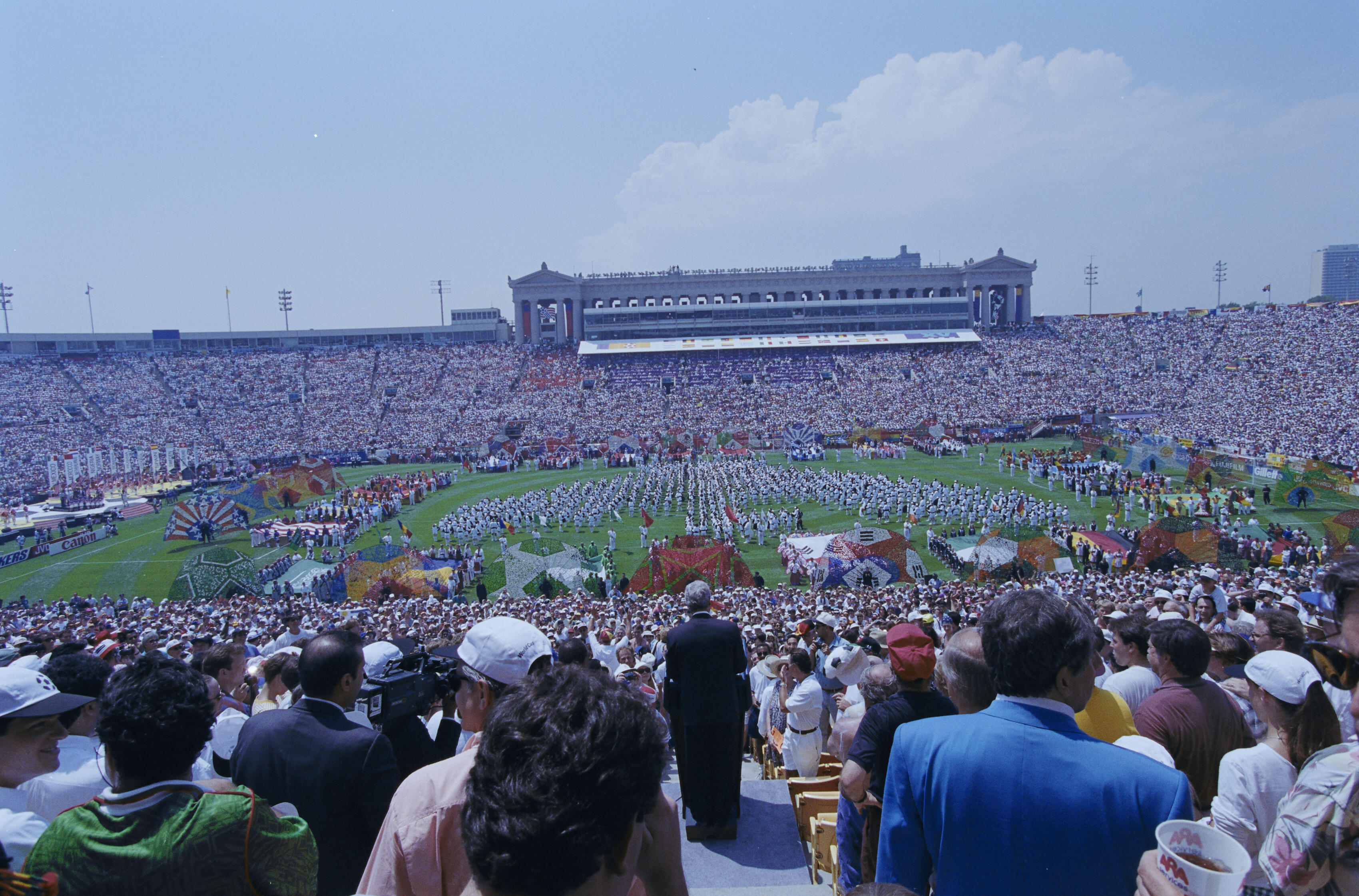 1994 FIFA World Cup opening ceremony at Soldier Field in Chicago. The stadium is filled to capacity with a dense crowd of spectators under a bright, partly cloudy sky. On the green field below, hundreds of performers in white uniforms are arranged in a large, circular formation, surrounded by vibrant displays of colorful fabrics and various international flags. In the foreground, the backs of spectators in the stands are visible, including a man in a dark suit standing prominently in the center aisle. 