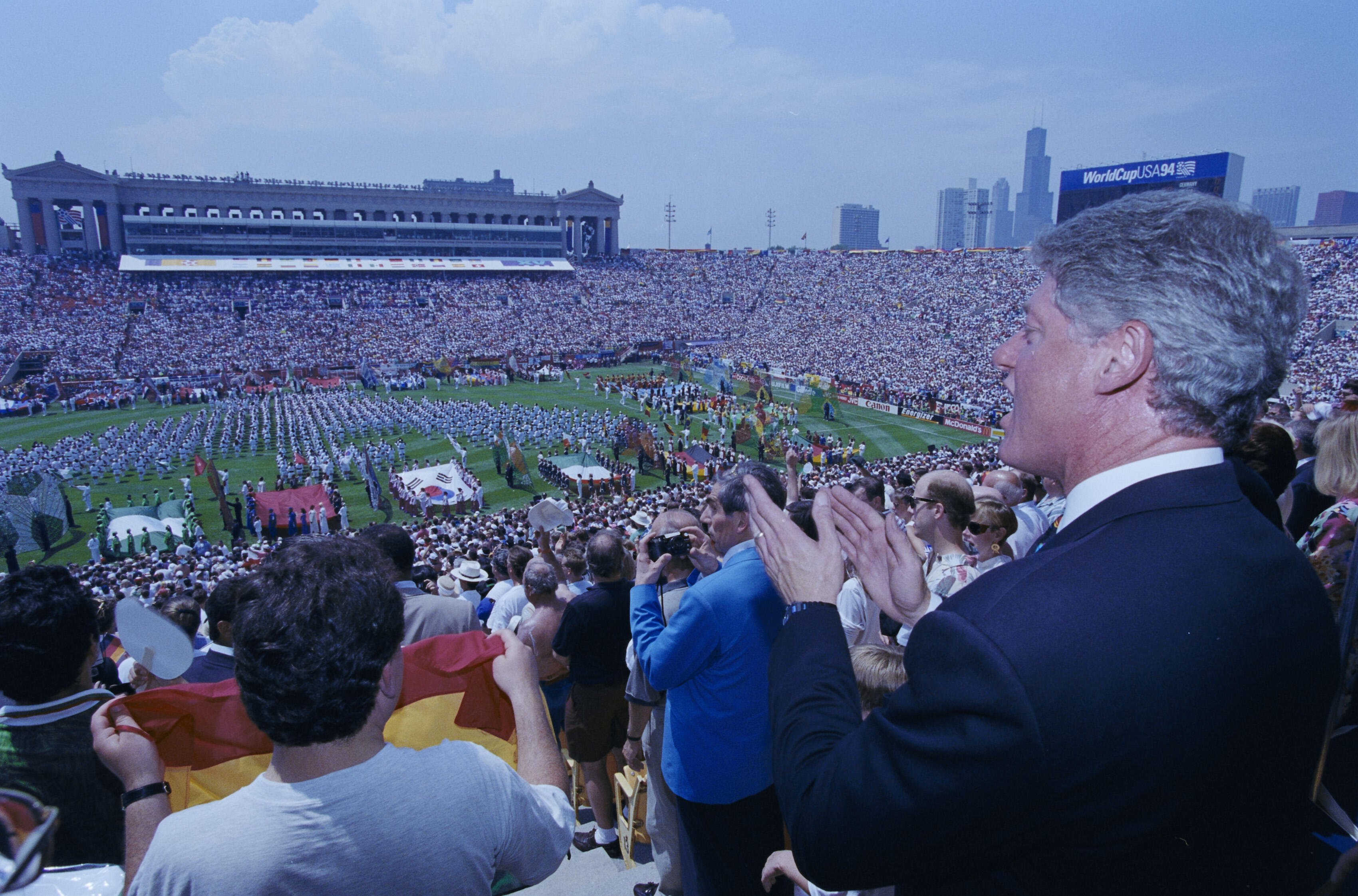 President Bill Clinton (on the right) looking out and applauding from the stands during the 1994 FIFA World Cup opening ceremony at Soldier Field. In the foreground, the backs and sides of various spectators are visible, including one person holding a small flag. The stadium is packed with a massive crowd. On the green field, hundreds of performers in white uniforms are organized into large formations, accompanied by colorful banners and flags, including the flag of South Korea. 