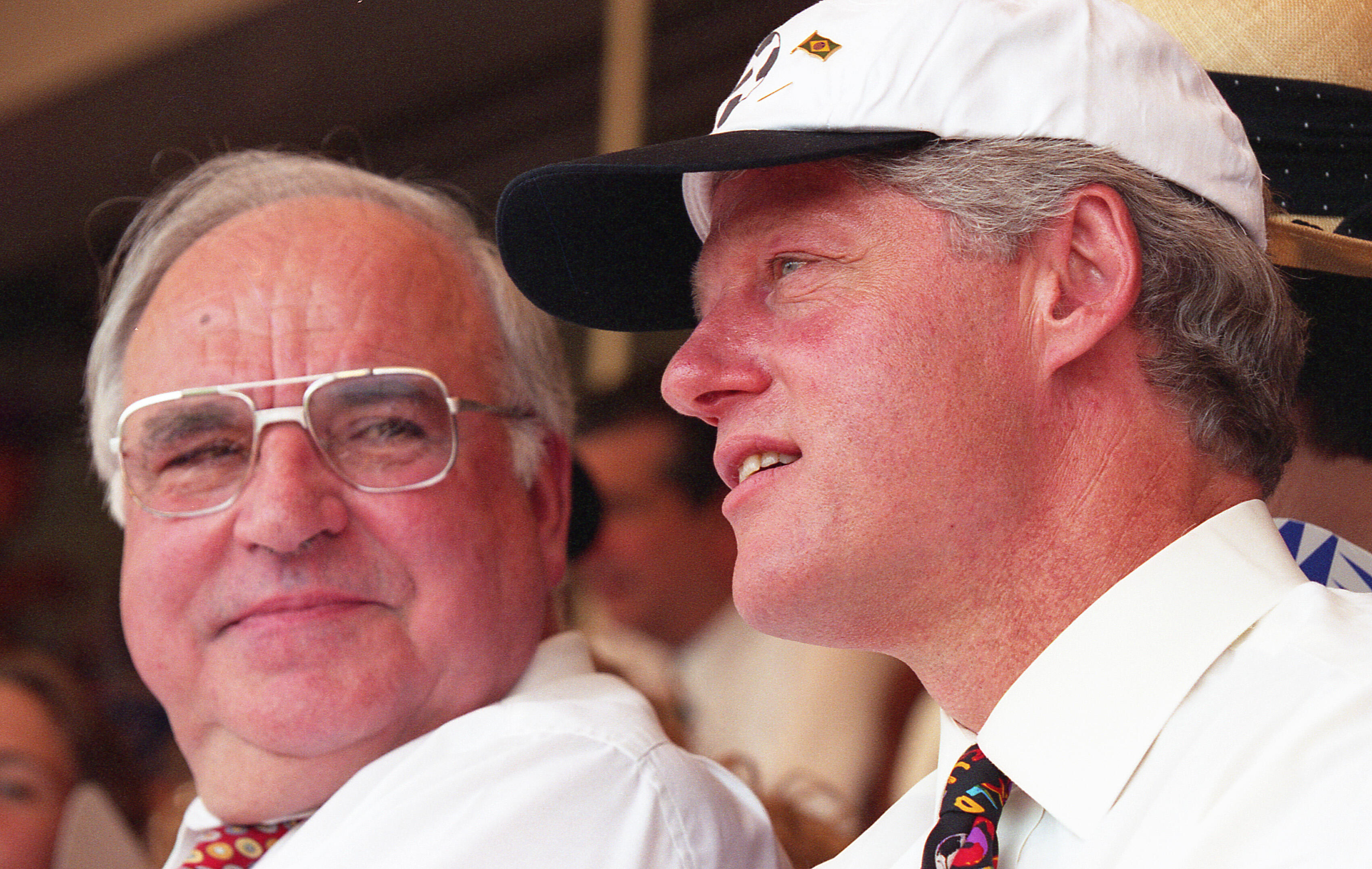 This close-up, eye-level photograph captures President Bill Clinton and German Chancellor Helmut Kohl watching the 1994 FIFA World Cup opening ceremony at Soldier Field. On the right, President Clinton is shown in profile, wearing a white shirt, a colorful patterned tie, and a white WorldCupUSA94 baseball cap with a black brim. To his left, Chancellor Kohl looks toward the field, wearing large, thin-framed glasses and a white shirt. Both men appear engaged and relaxed during the festivities.
