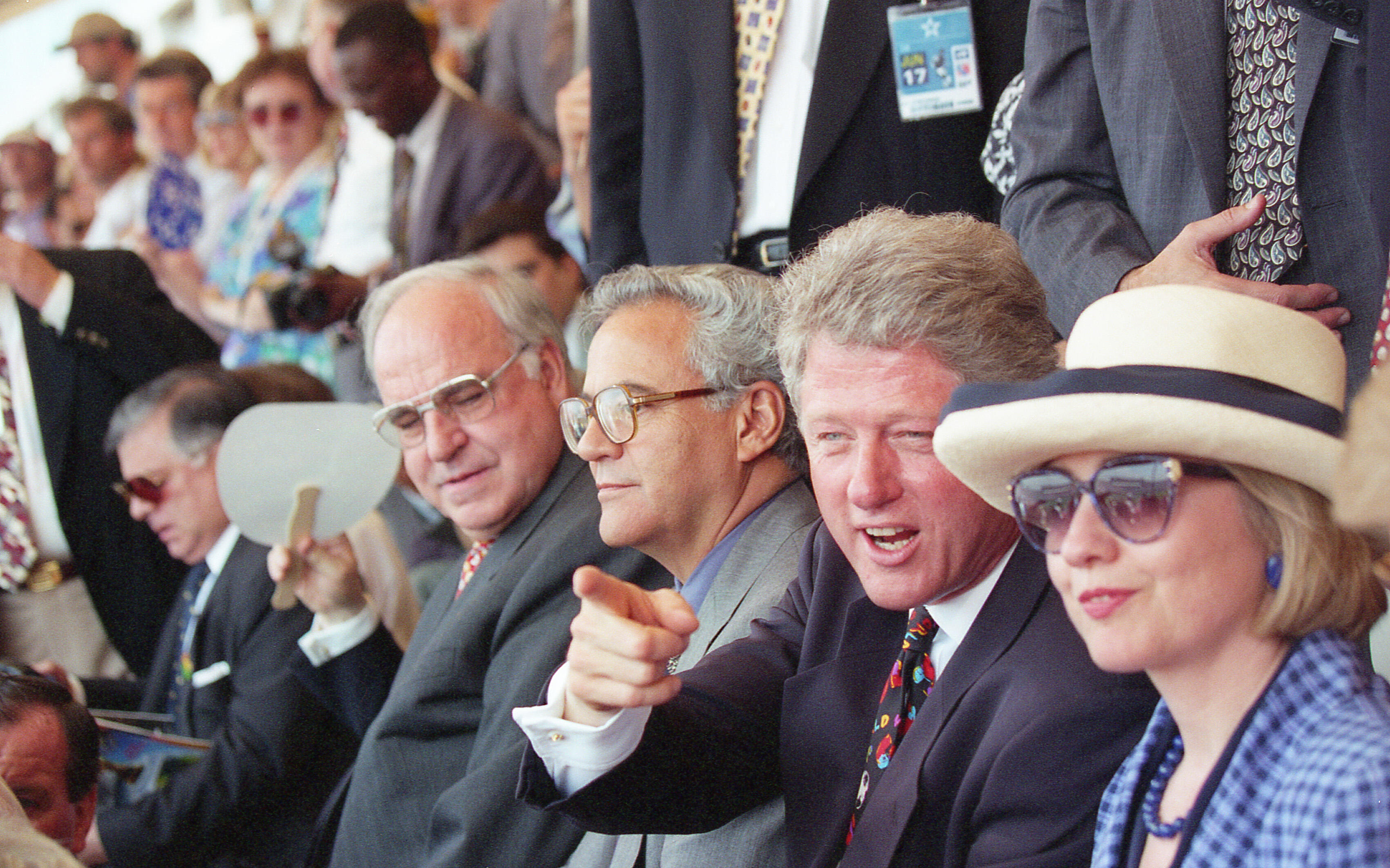 President Bill Clinton is shown speaking and gesturing toward the field while wearing a dark suit and a colorful patterned tie.Hillary Rodham Clinton: The First Lady sits next to him, wearing a blue-and-white checkered blazer and a wide-brimmed cream hat.Helmut Kohl: The former German Chancellor is seated to the President's right (on the left side of the frame), wearing large, thin-framed glasses.
