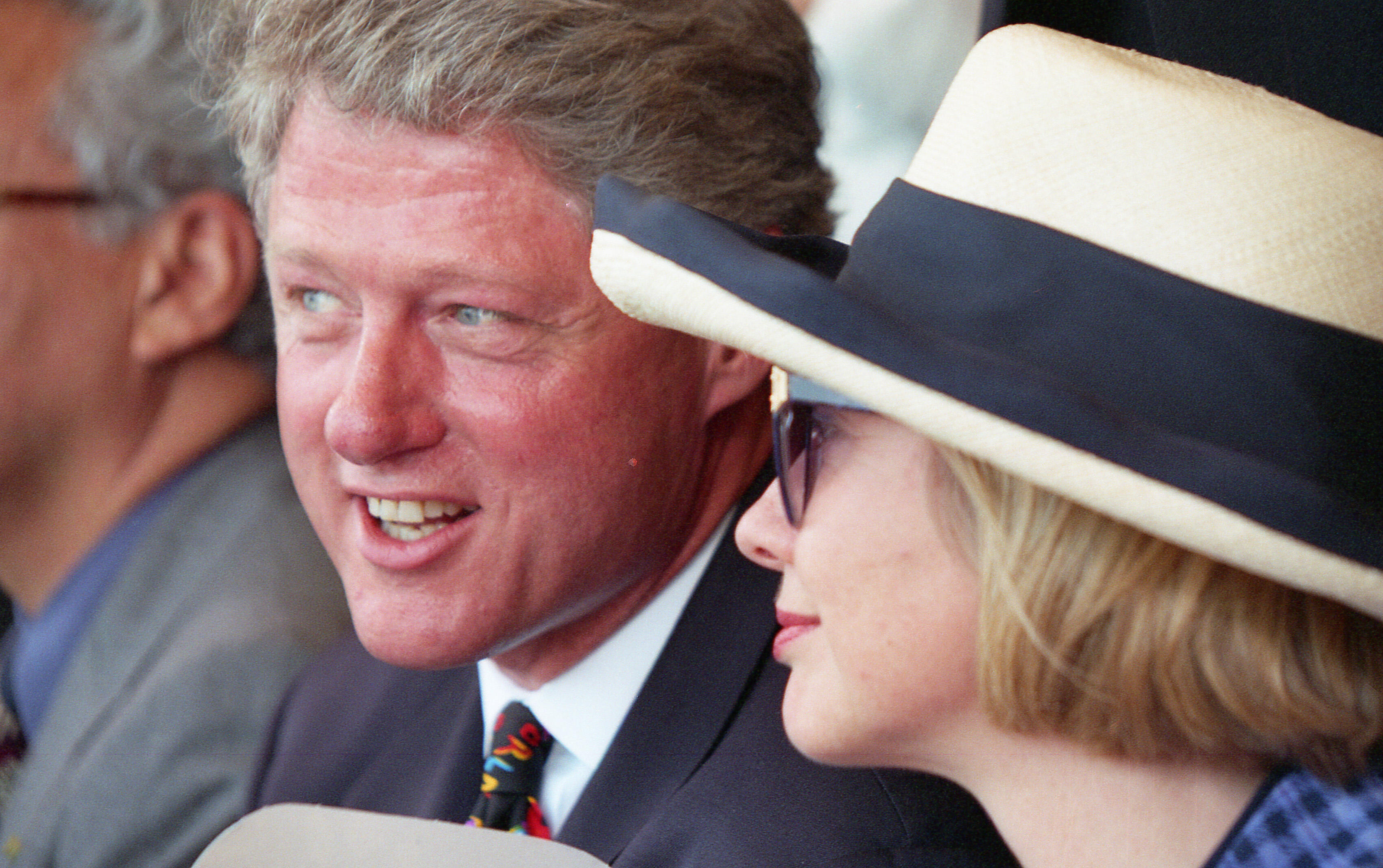 On the left, President Clinton is shown in a three-quarter view, smiling and looking toward the field. He wears a dark suit and a colorful patterned tie. To his right, Hillary Clinton is seen in profile, wearing a blue checkered blazer and a wide-brimmed cream hat with a black band. A third person, partially visible on the far left, wears glasses and a gray suit. The lighting is bright and natural, typical of an outdoor afternoon event.
