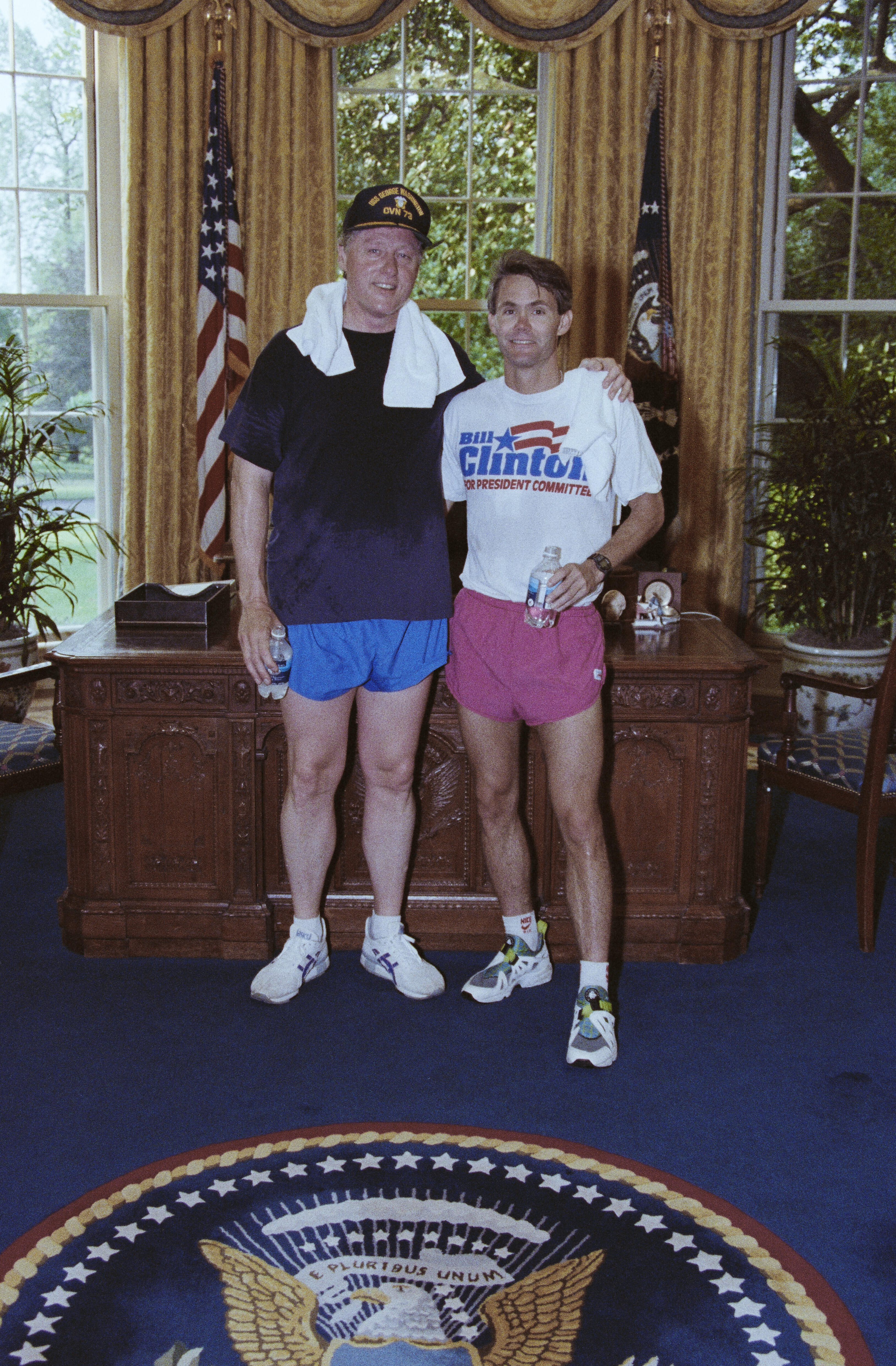 President Clinton and Billy Webster pose for a photograph in the Oval Office after a jog along the Potomac River. President Clinton has a white towel wrapped around his neck. Both men stand. President Clinton his hand on Webster's shoulder. Webster holds a bottle of water in his hand.