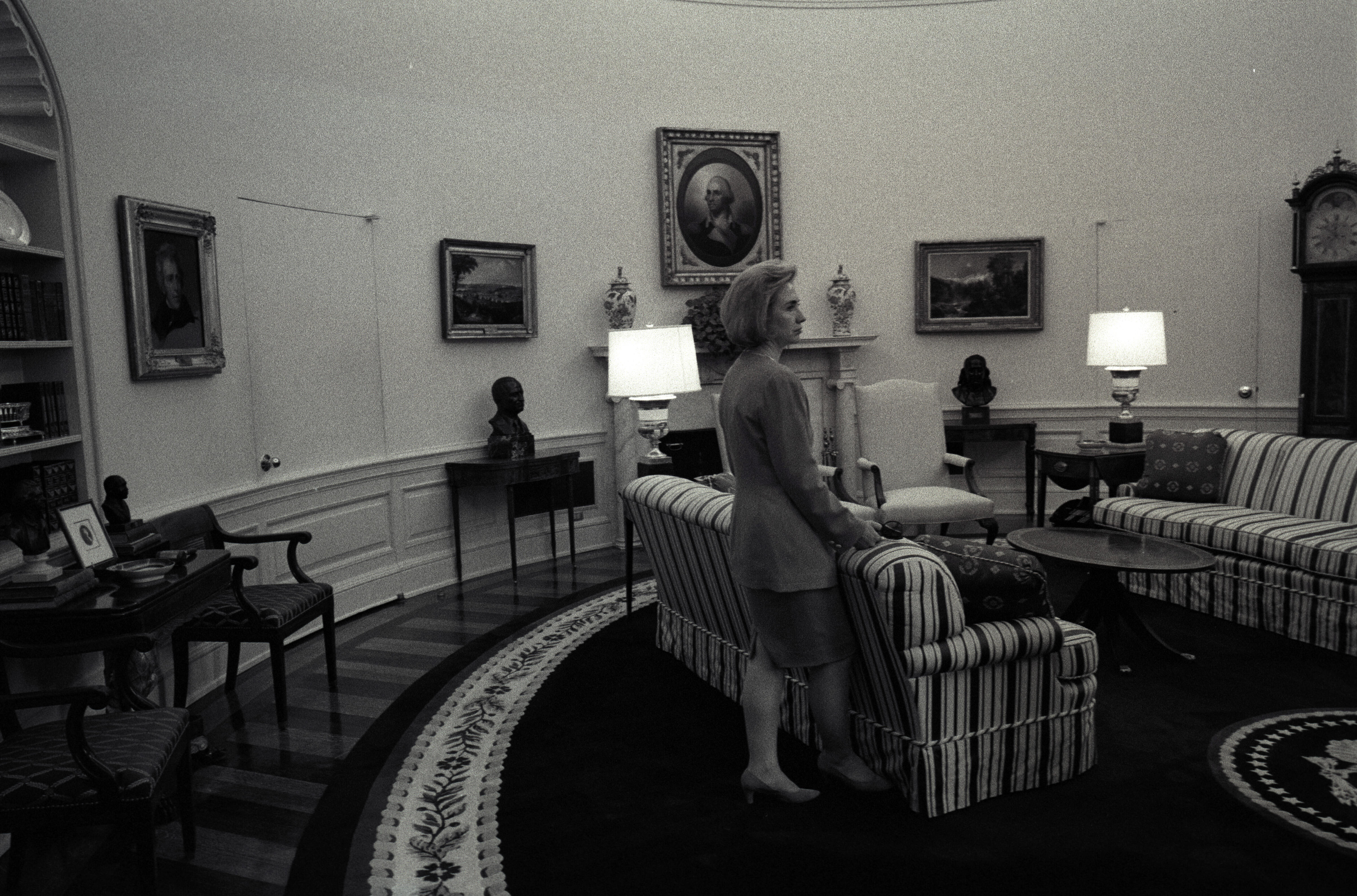 Black and white photograph. First Lady Hillary Rodham Clinton standing alone next to a striped sofa in the Oval Office