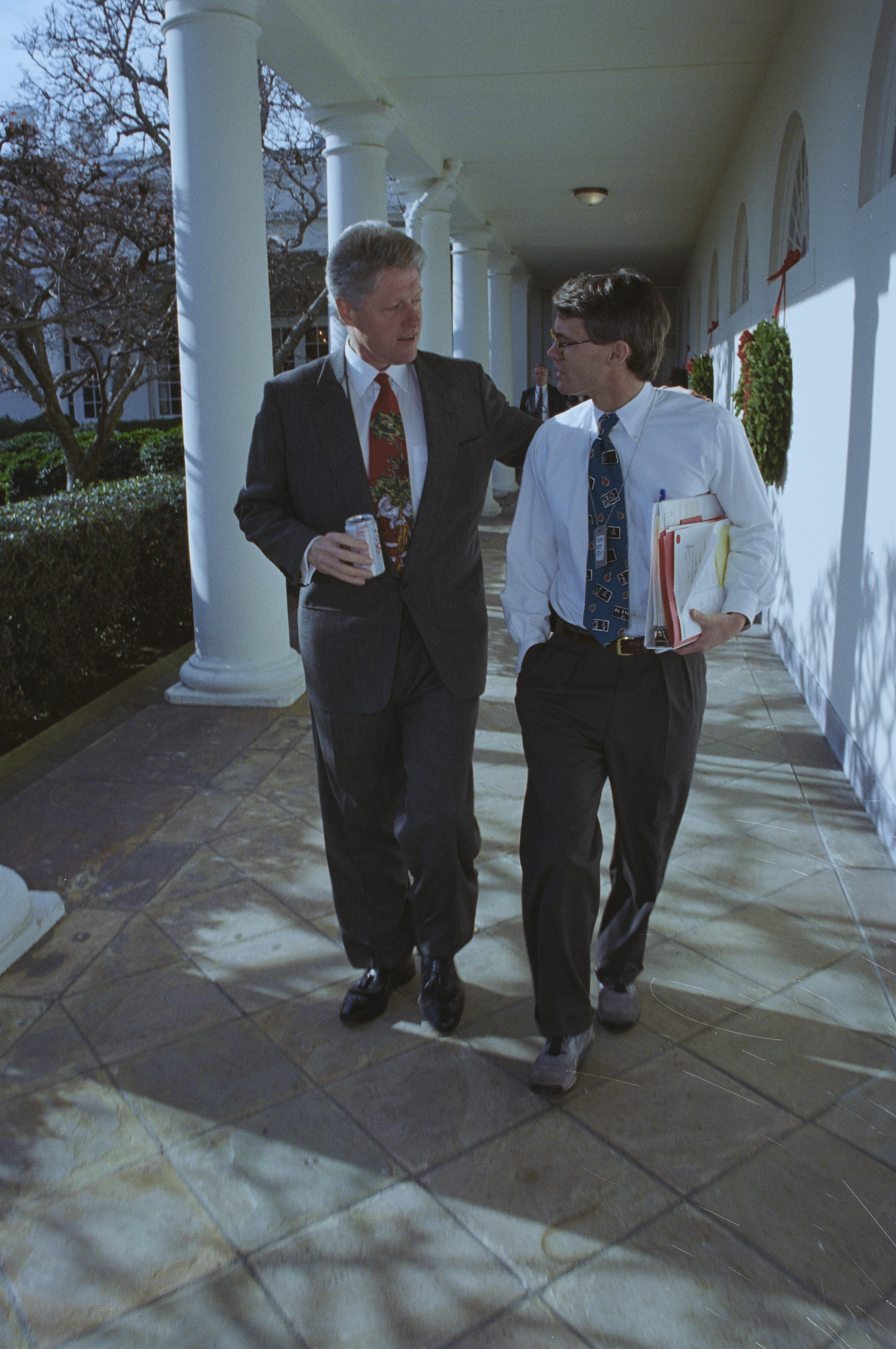 President Clinton and Billy Webster walk alone down the colonnade. Webster holds files in his hand. President Clinton holds a drink in his hand as they walk and talk.
