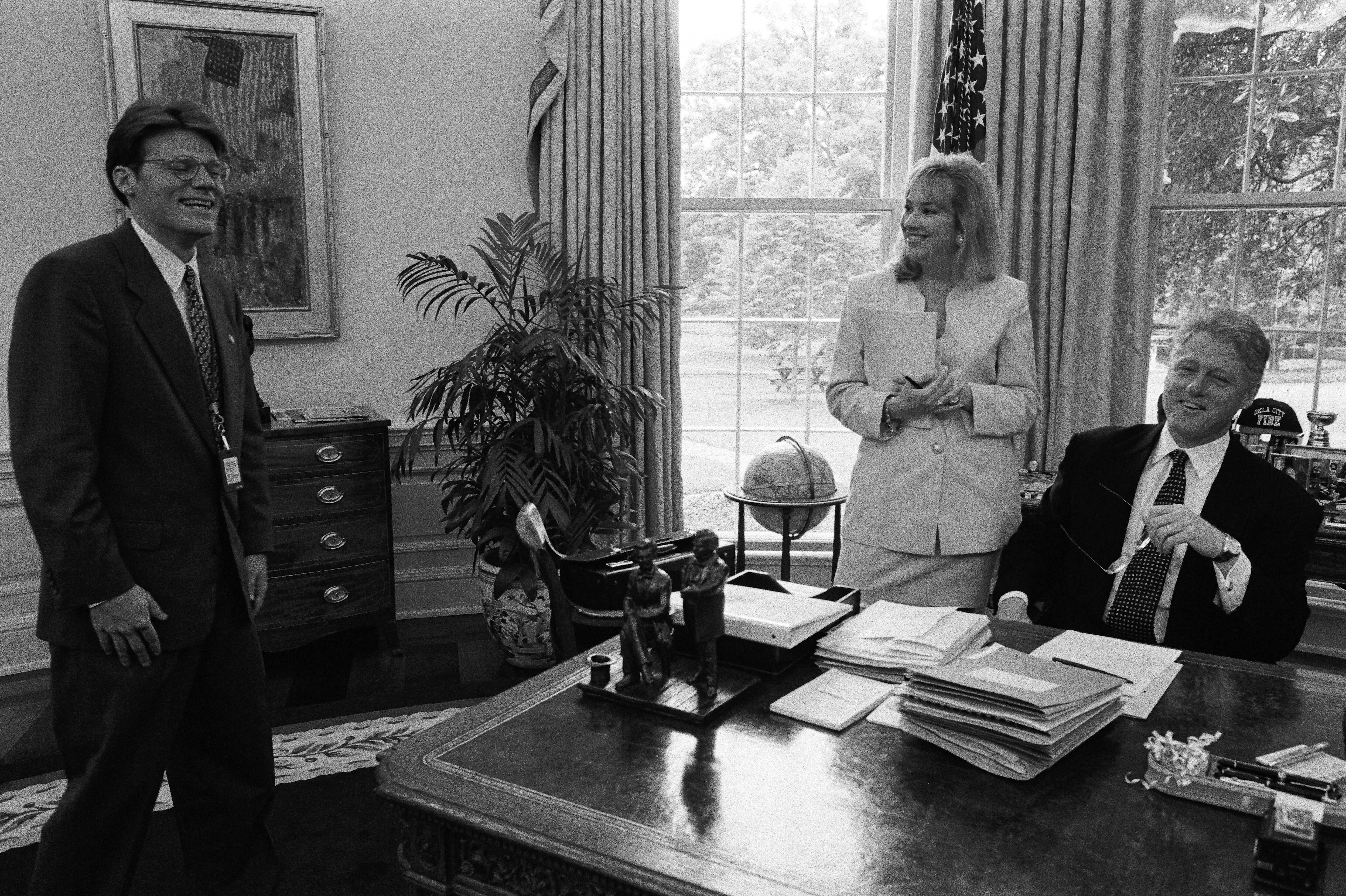 Black and white photograph. President Clinton meets with Director of the Office of Scheduling and Advance, Stephanie Streett, and President's Aide, Stephen Goodin in the White House Oval Office. Goodin and Streett are standing, President Clinton sits behind his desk. All are smiling.