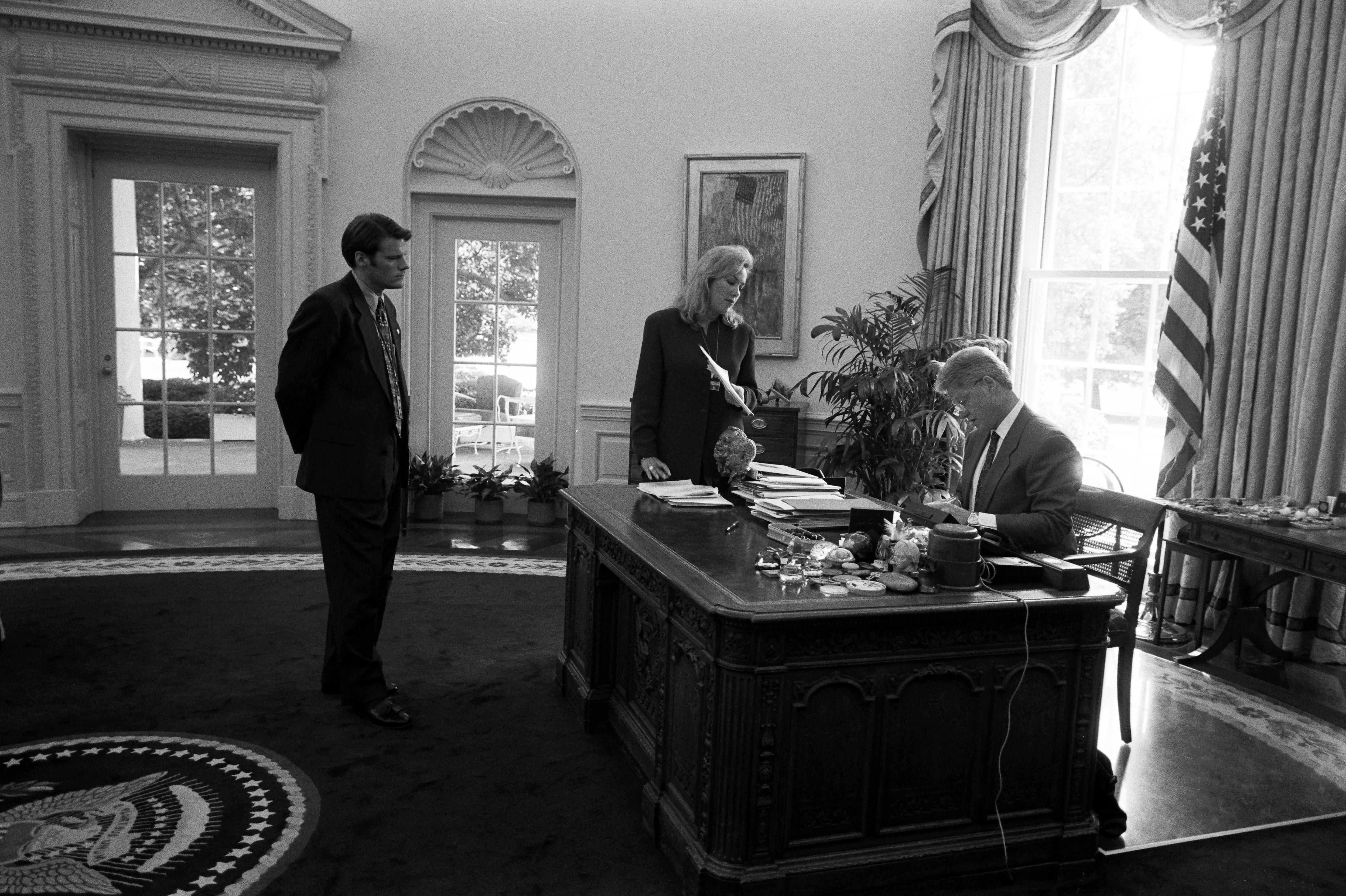President Clinton meets with Director of the Office of Scheduling and Advance, Stephanie Streett, and President's Aide, Stephen Goodin in the White House Oval Office. Streett and Goodin stand in front of the President's desk. President Clinton sits, looking at a document on his desk 
