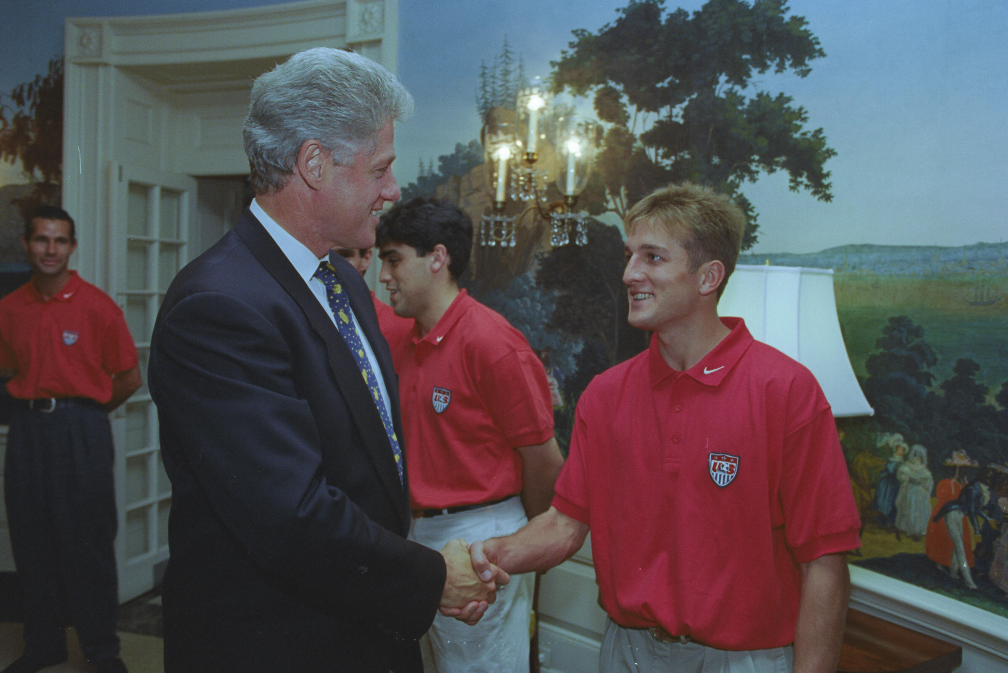 A medium shot capturing President Bill Clinton on the left, smiling as he shakes hands with a blonde man wearing a red U.S. soccer team polo shirt. In the background, two other team members in identical red shirts look on, with one standing partially behind the President. The room is decorated with a scenic nature mural on the wall and an elegant crystal wall sconce light fixture.
