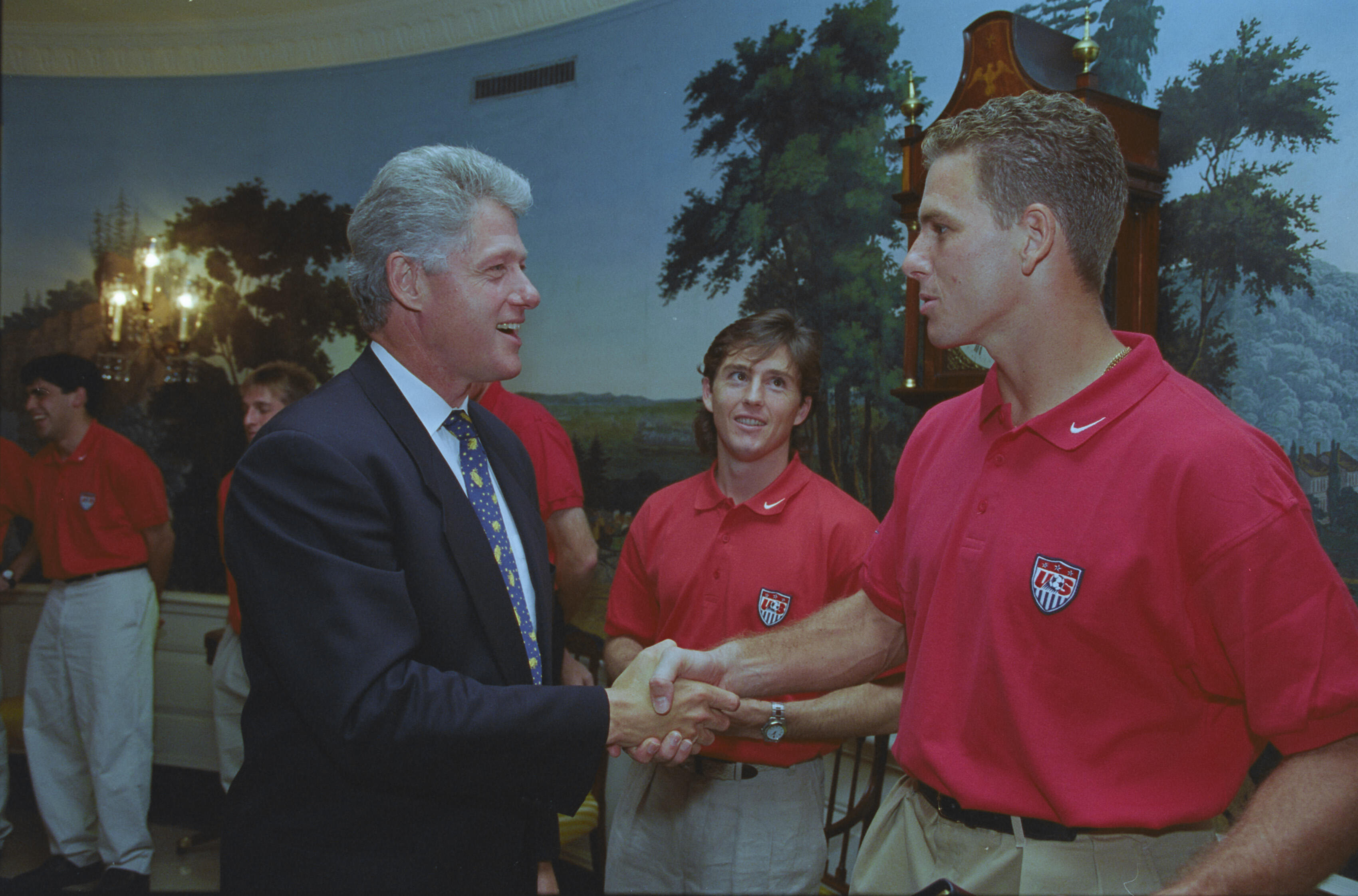 A medium shot capturing President Bill Clinton on the left, smiling as he shakes hands with a tall man with short blond hair wearing a red U.S. soccer team polo shirt. In the background, another team member with longer brown hair stands between them, looking toward the president. The setting features a scenic nature mural covering the wall, an elaborate wooden grandfather clock to the right, and an ornate lit wall sconce on the far left.