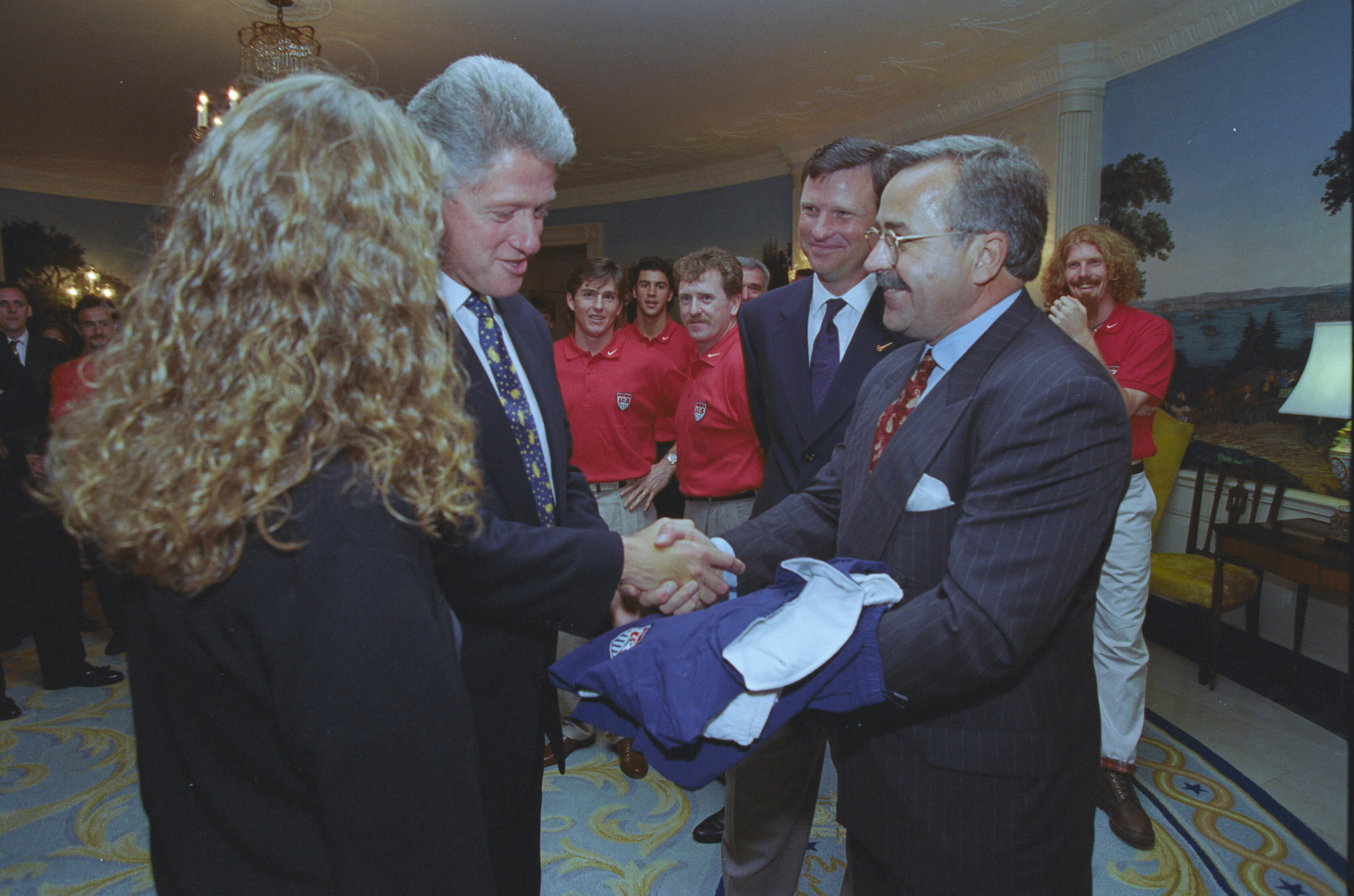 A medium shot capturing President Bill Clinton shaking hands with a man wearing a pinstripe suit, mustache, and glasses. The man holds a folded blue and white jacket or jersey under his arm during the handshake. In the left foreground, Chelsea Clinton is seen from behind, showing her long curly blonde hair. In the background, several members of the U.S. Men's National Soccer Team look on in red polo shirts, alongside another official in a dark blue suit. 