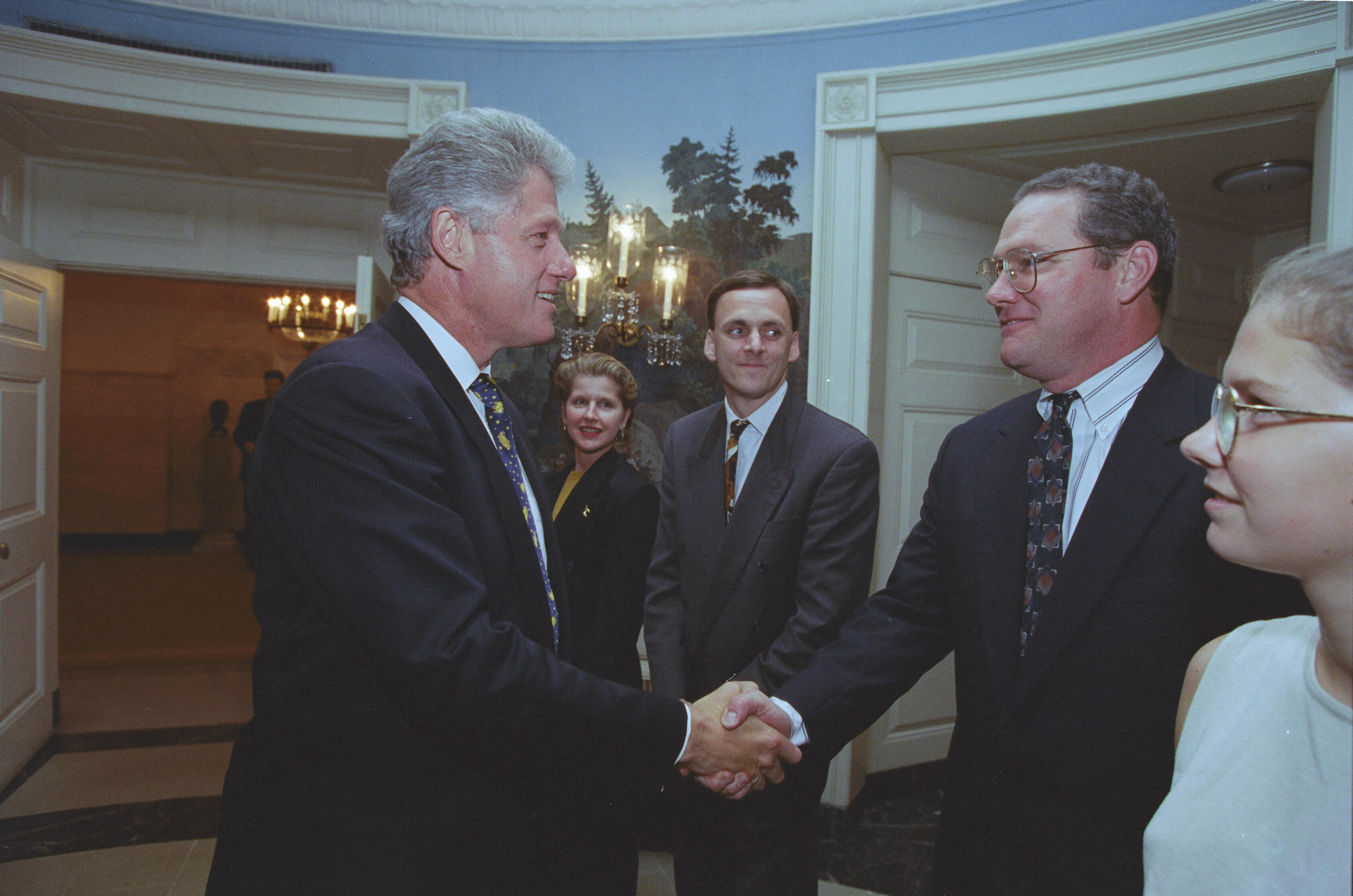 A photograph captures President Bill Clinton shaking hands with a man wearing glasses and a dark suit inside a White House room. Clinton stands on the left, smiling as he grips the man's hand. To the right, a young woman wearing glasses is seen in partial profile, looking toward the interaction. In the background, a woman with blonde hair and the man from the previous photograph stand smiling. They are positioned in front of a wall with scenic wallpaper and a decorative wall sconce.