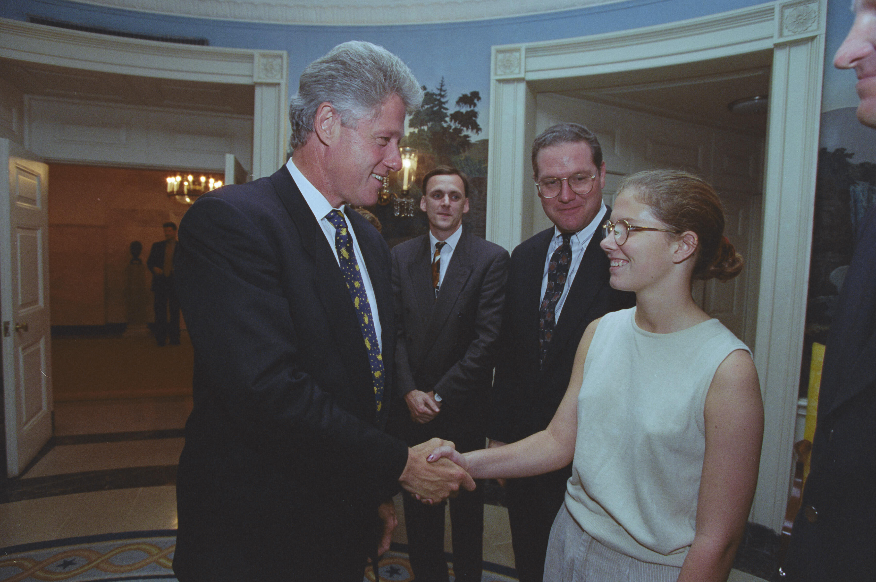 A photograph captures President Bill Clinton smiling as he shakes hands with a young woman wearing glasses and a light-colored sleeveless top. Clinton stands on the left in a dark suit and patterned tie. The young woman stands on the right, looking back at him with a smile. In the background between them, two men in suits look on—the man on the right wears glasses and smiles, while the man in the center watches the handshake. They are all gathered in a White House room featuring scenic wallpaper.