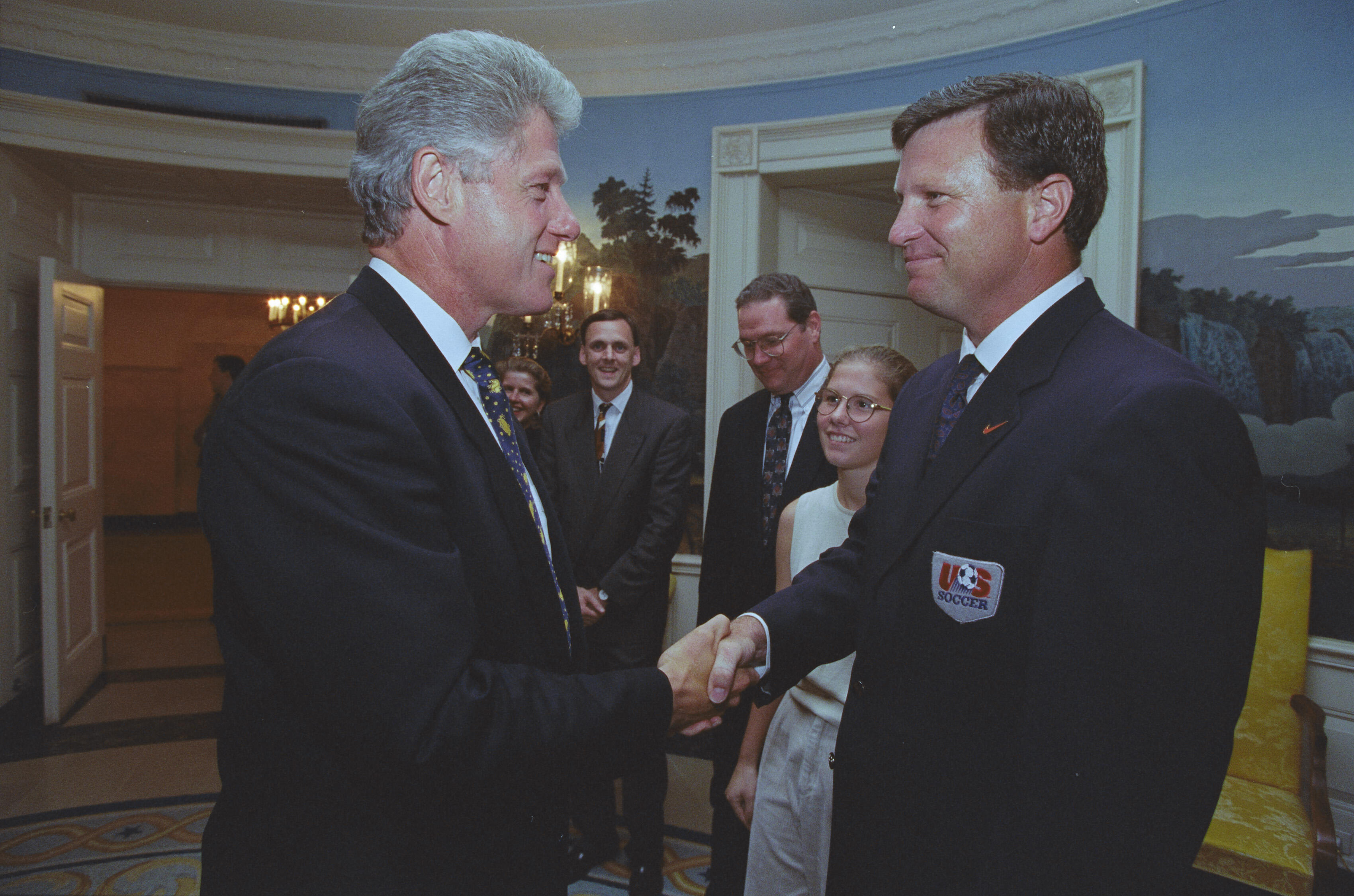 A medium shot capturing President Bill Clinton on the left, smiling as he shakes hands with a man wearing a dark blazer with a U.S. Soccer patch on the pocket. In the background between them, Chelsea Clinton stands smiling, wearing glasses and a sleeveless top, alongside two men in dark suits and patterned ties. The room features a scenic nature mural on the wall, an open doorway revealing a lit chandelier in an adjacent hallway, and a yellow upholstered chair visible on the far right.