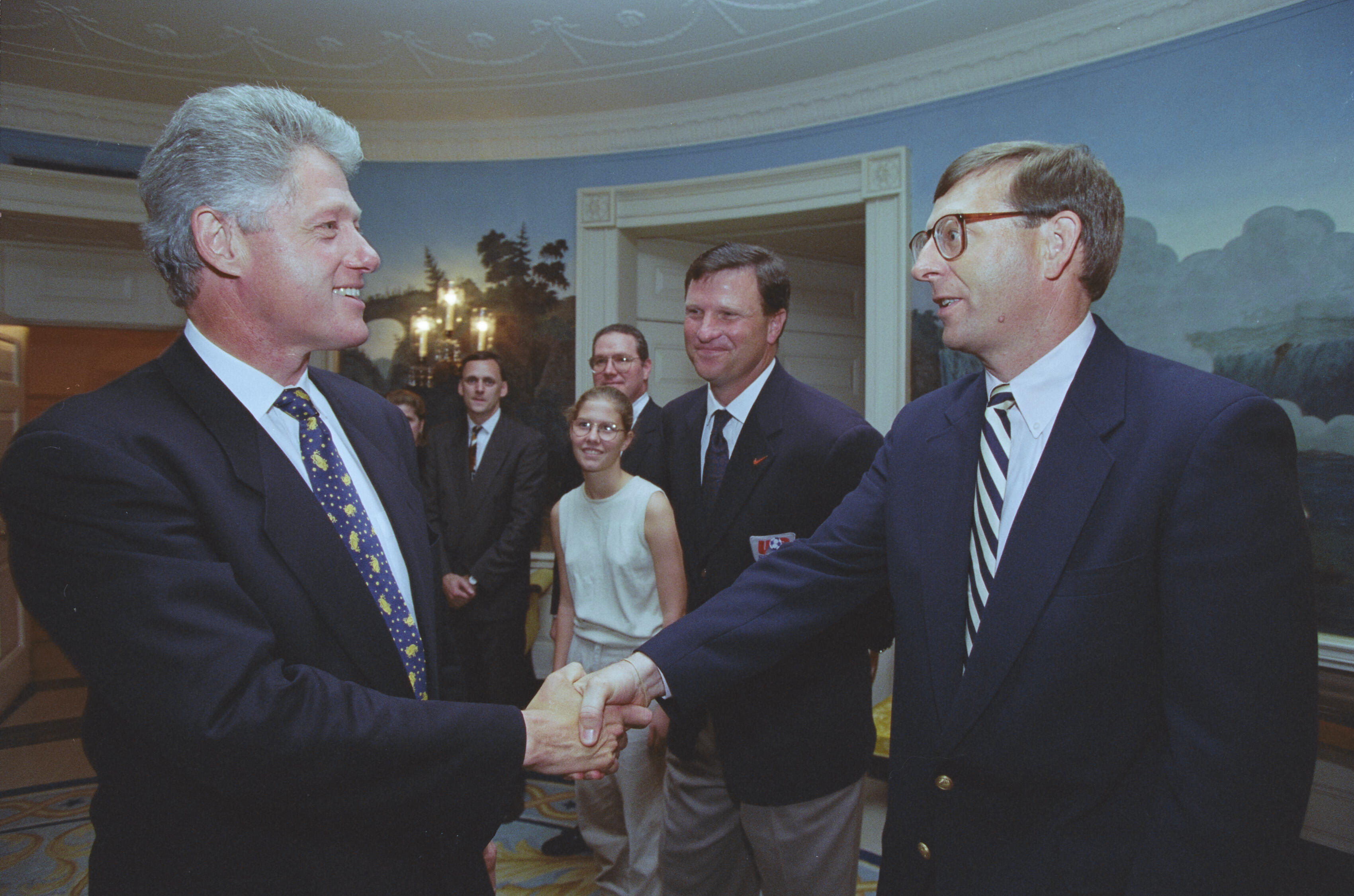 A photograph captures President Bill Clinton smiling as he shakes hands with a man wearing glasses and a striped tie. Clinton stands on the left in a dark suit with a patterned blue tie. The man stands on the right, looking toward Clinton while shaking his hand. In the background, several individuals look on, including a man in a dark blazer with a red logo badge, a young woman wearing glasses and a sleeveless top, and two other men in suits. They are gathered in a White House room with light blue walls.