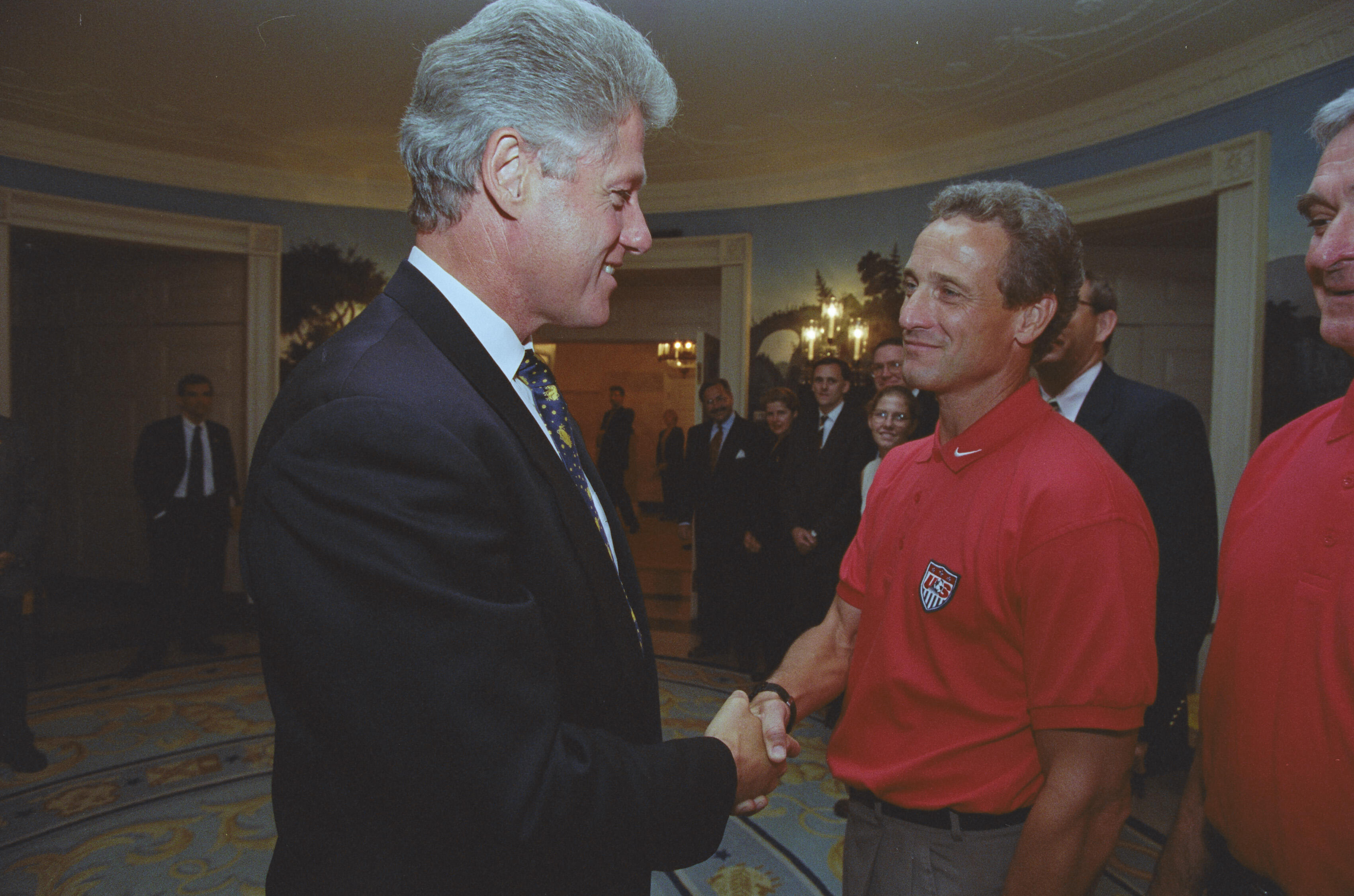 A photograph captures President Bill Clinton smiling as he shakes hands with a man wearing a red polo shirt. Clinton stands on the left in a dark suit with a patterned tie. The man stands on the right, looking back at Clinton with a slight smile; his red shirt features a USA crest on the left chest. In the background, several people look on from across a large room with blue walls, patterned carpeting, and scenic wallpaper.