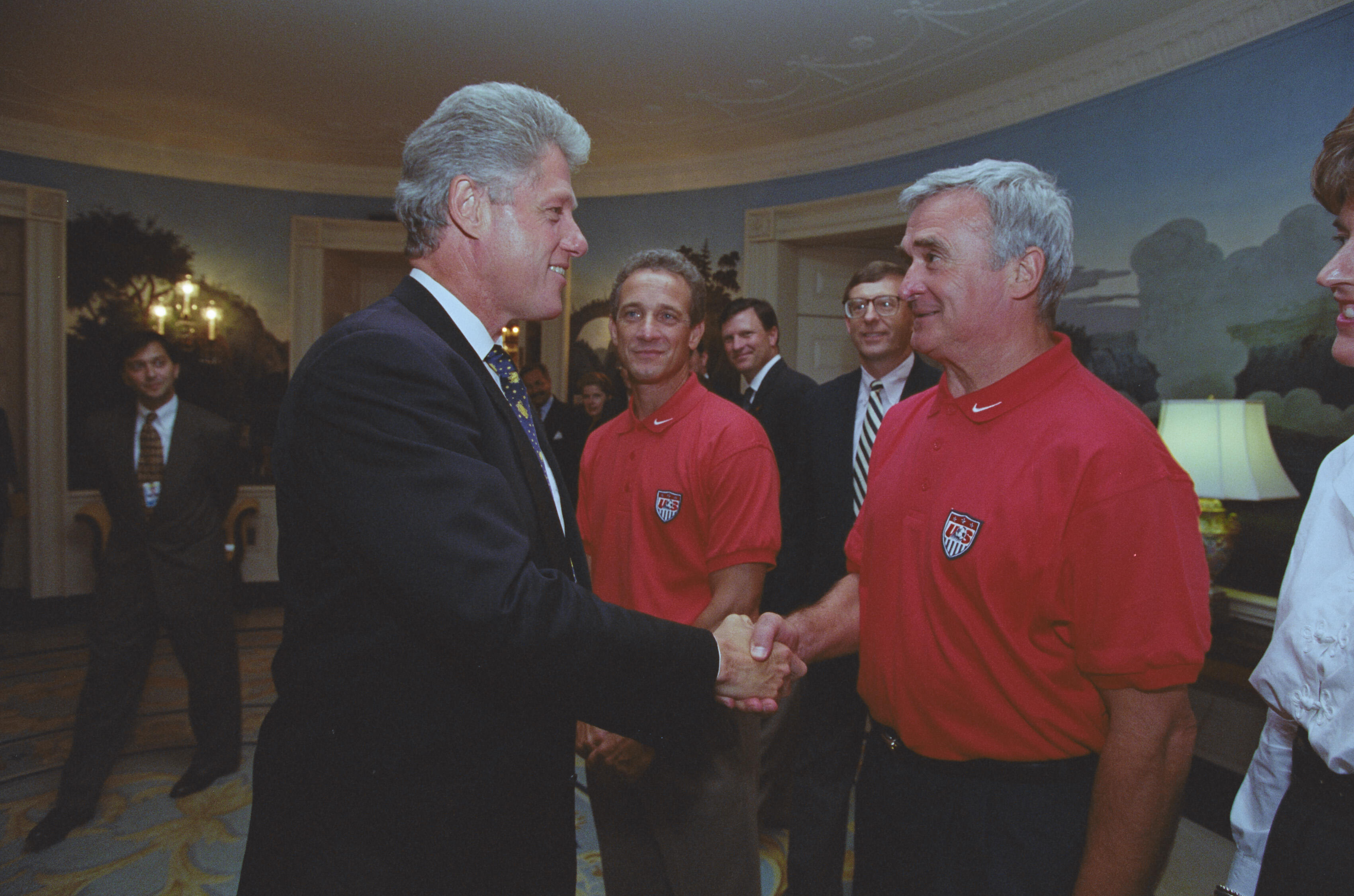 A photograph captures President Bill Clinton smiling as he shakes hands with an older man with grey hair wearing a red polo shirt. Clinton stands on the left in a dark suit. The man stands on the right, looking toward Clinton; his red polo features a USA crest on the left chest. In the background between them, the man from the previous photograph stands watching in an identical red polo shirt. Other onlookers in suits stand behind them in the White House room, which features scenic wallpaper.