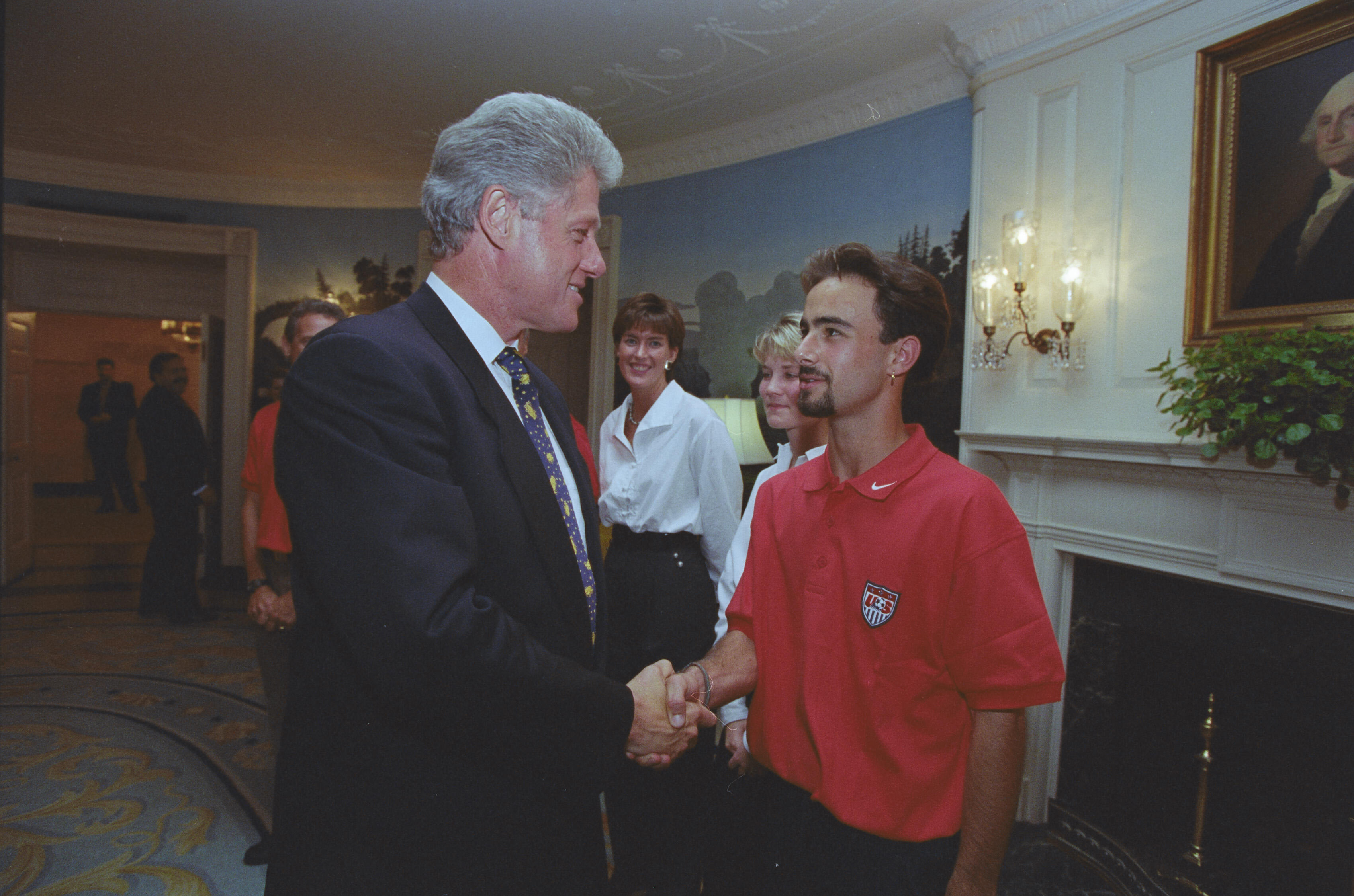 A photograph of President Bill Clinton shaking hands with a young man in the White House. Clinton, seen in profile wearing a dark suit and patterned tie, smiles as he grasps the hand of the man, who wears a red polo shirt featuring a U.S. Soccer logo. In the background, several onlookers smile, and a portrait of George Washington hangs above a nearby fireplace.